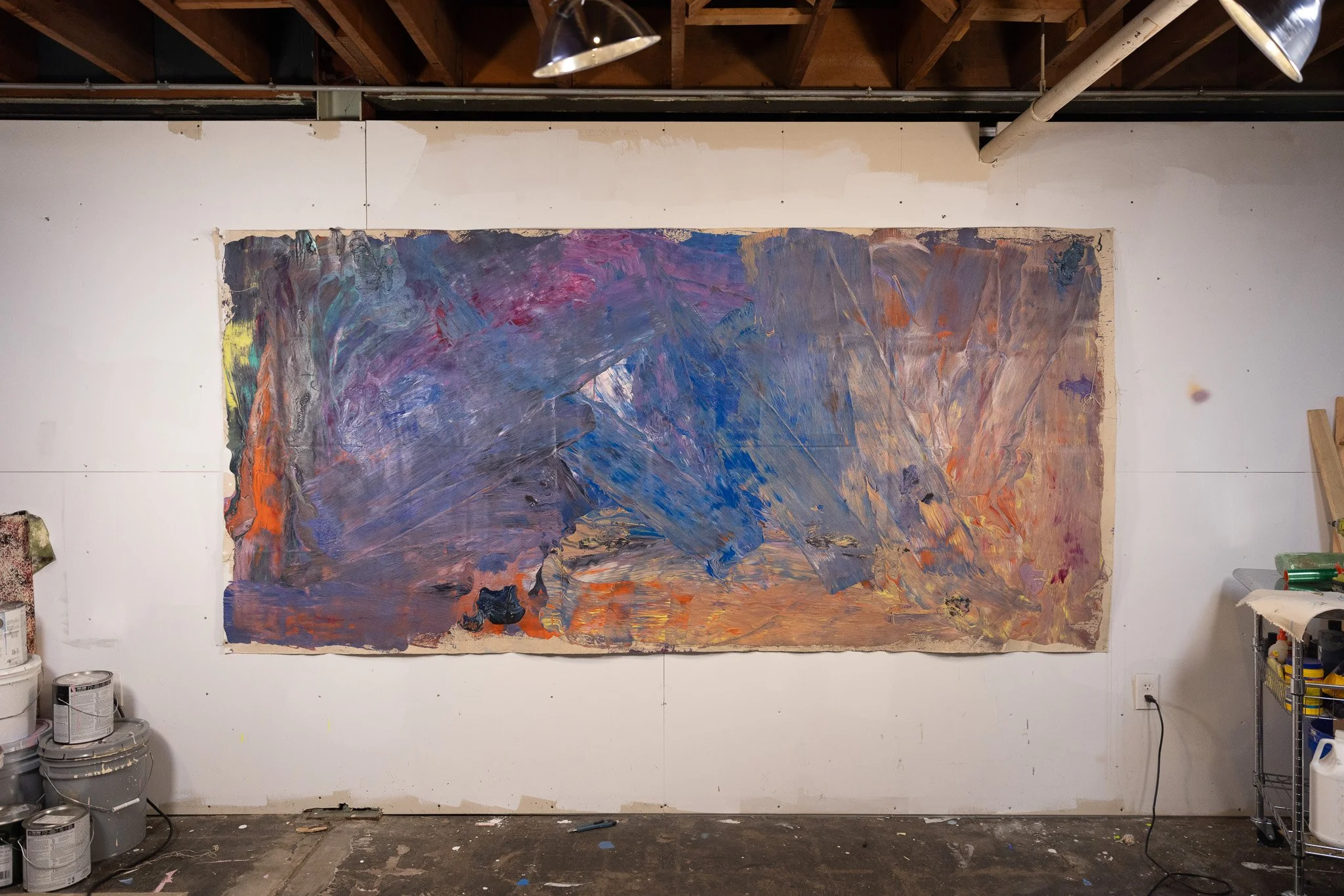 Large abstract painting with vibrant colors on a wall in an art studio, surrounded by paint cans and art supplies.
