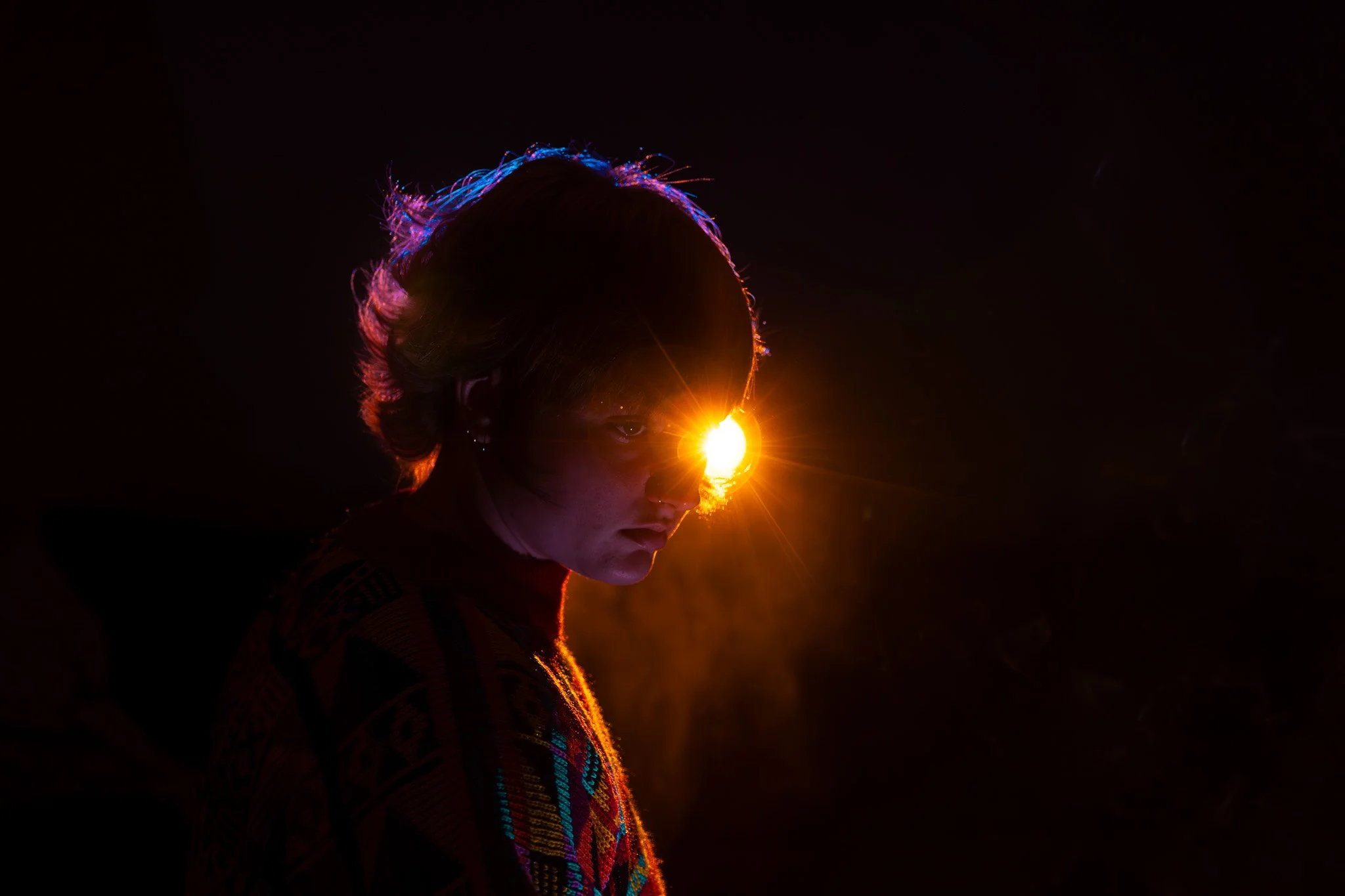 Person with short hair looking over shoulder against dark background, illuminated by bright orange light source near face.