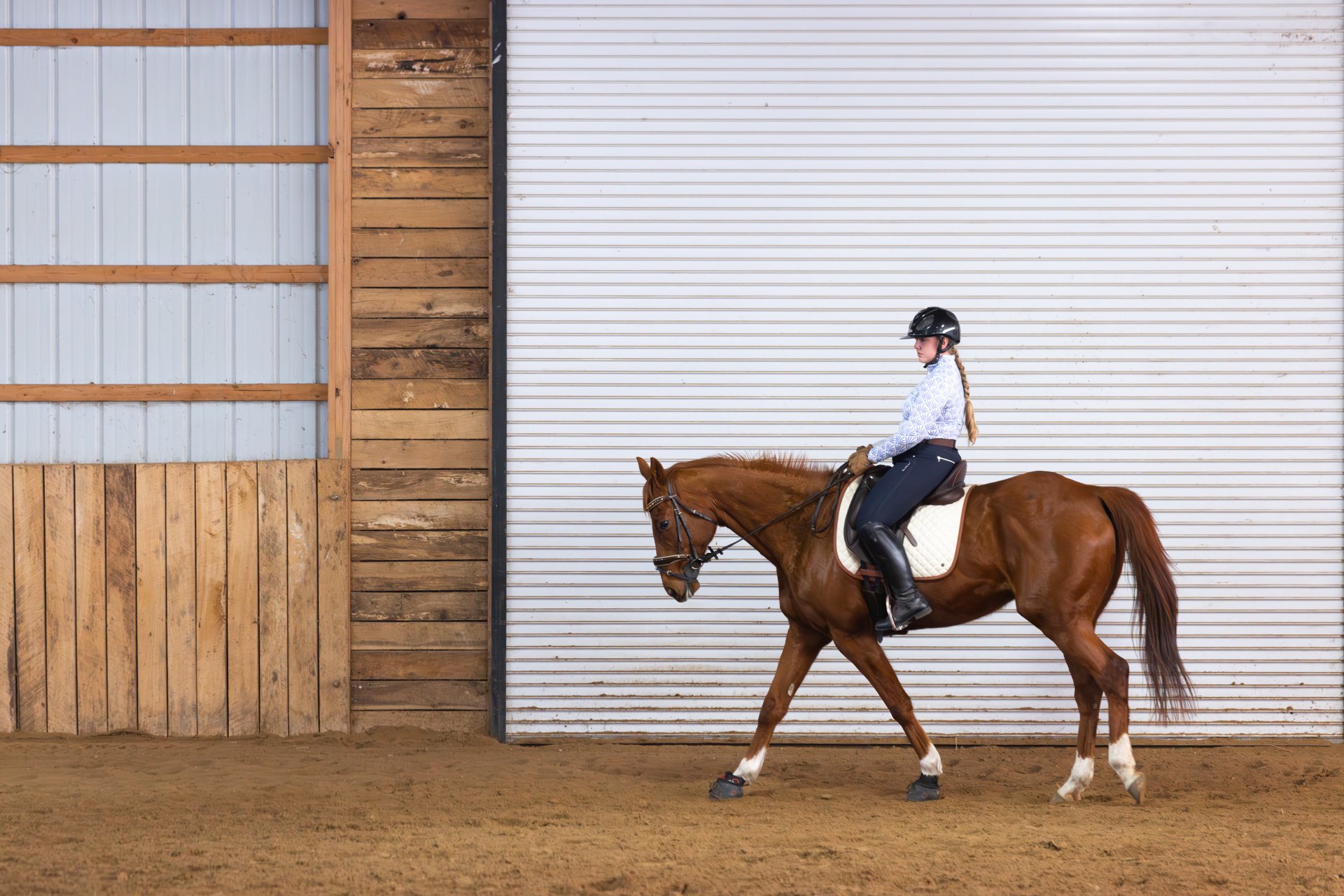A woman riding a brown horse inside an indoor riding arena with wooden and metal walls.