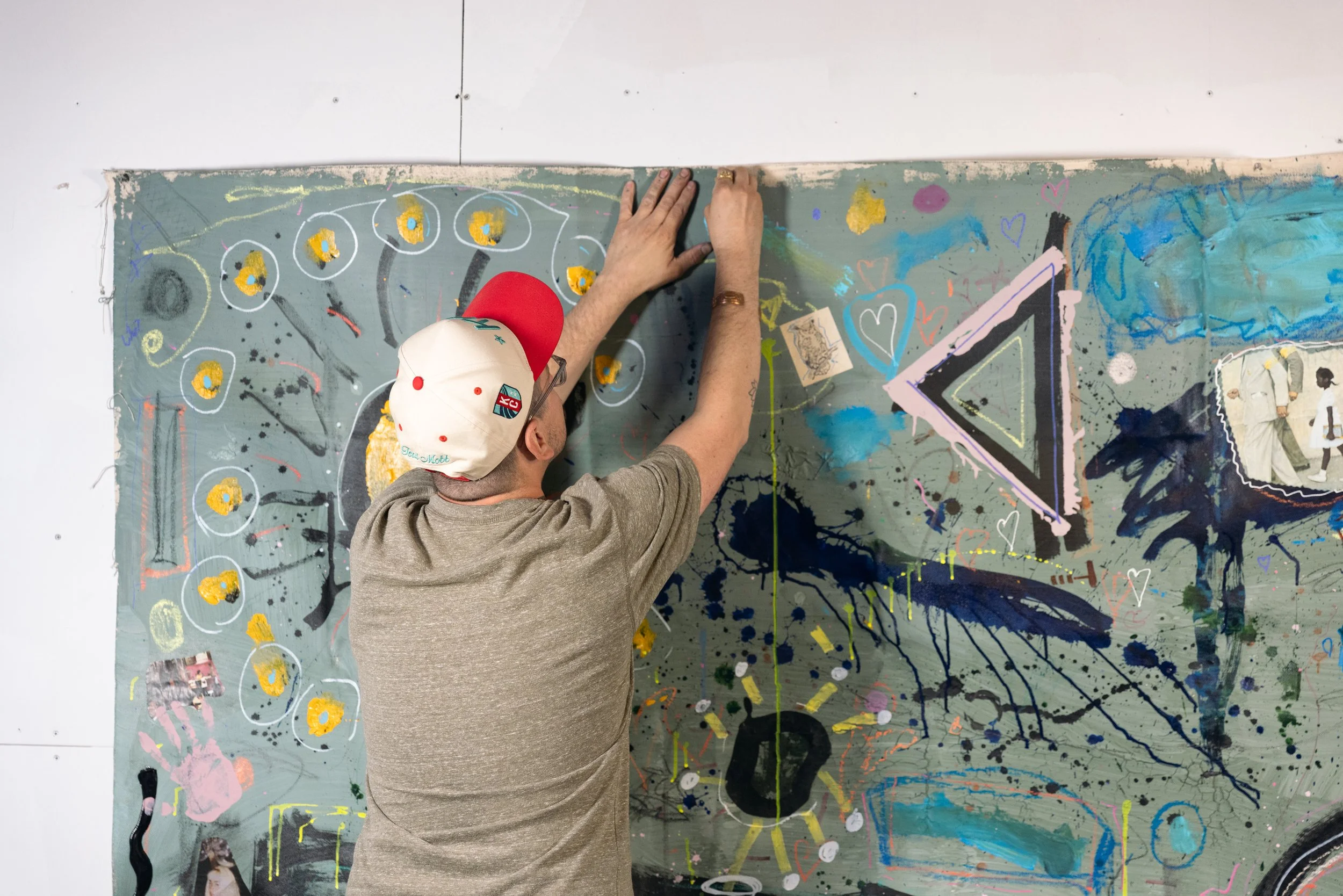 A man with a beige baseball cap and glasses creating modern abstract art on a large painted canvas with various colors, shapes, and symbols, including hearts and a triangle.