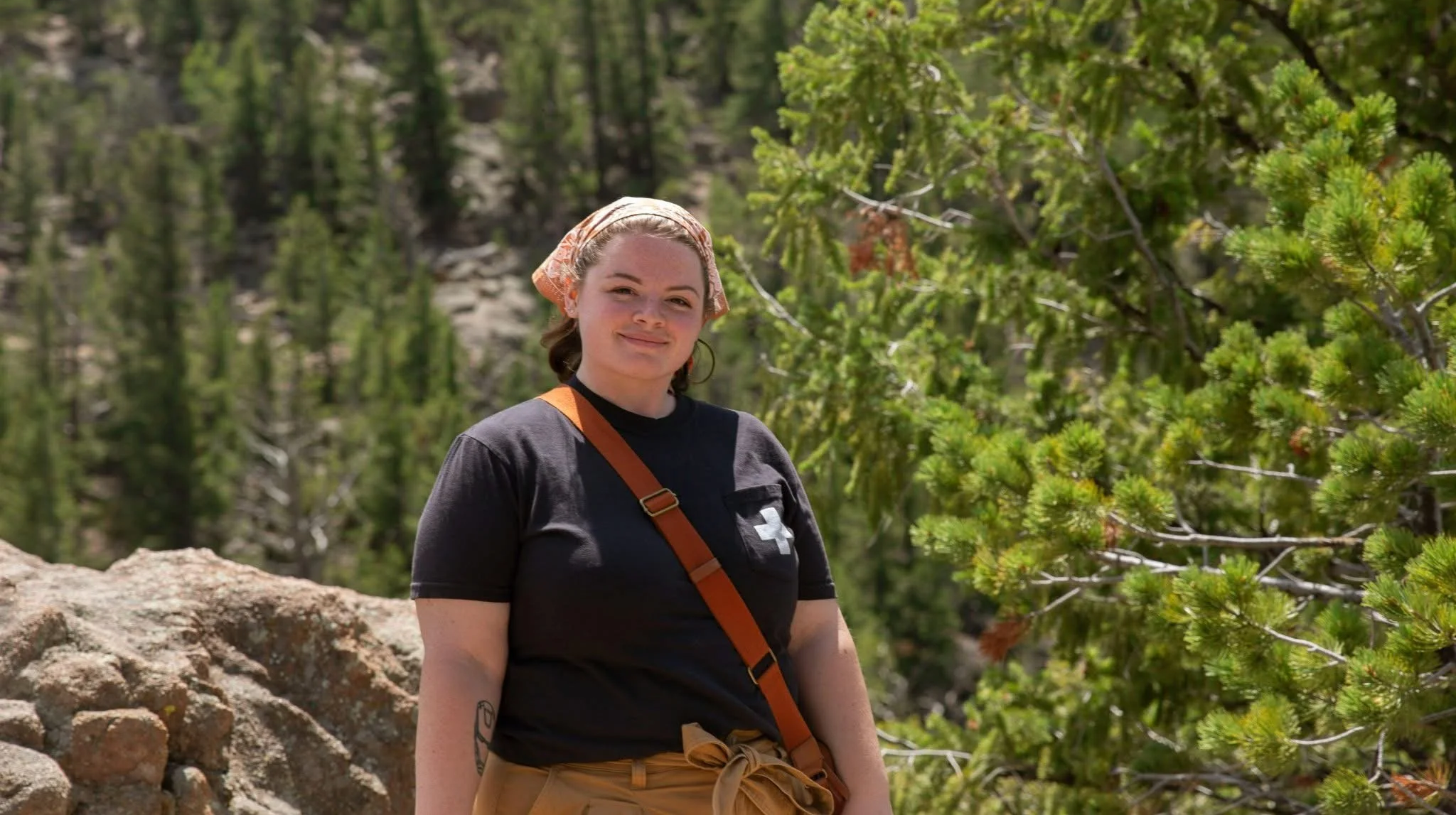 A woman with a bandana on her head, wearing a black t-shirt with a white cross on it, and khaki pants, standing outdoors in a forested area with green trees and rocks.