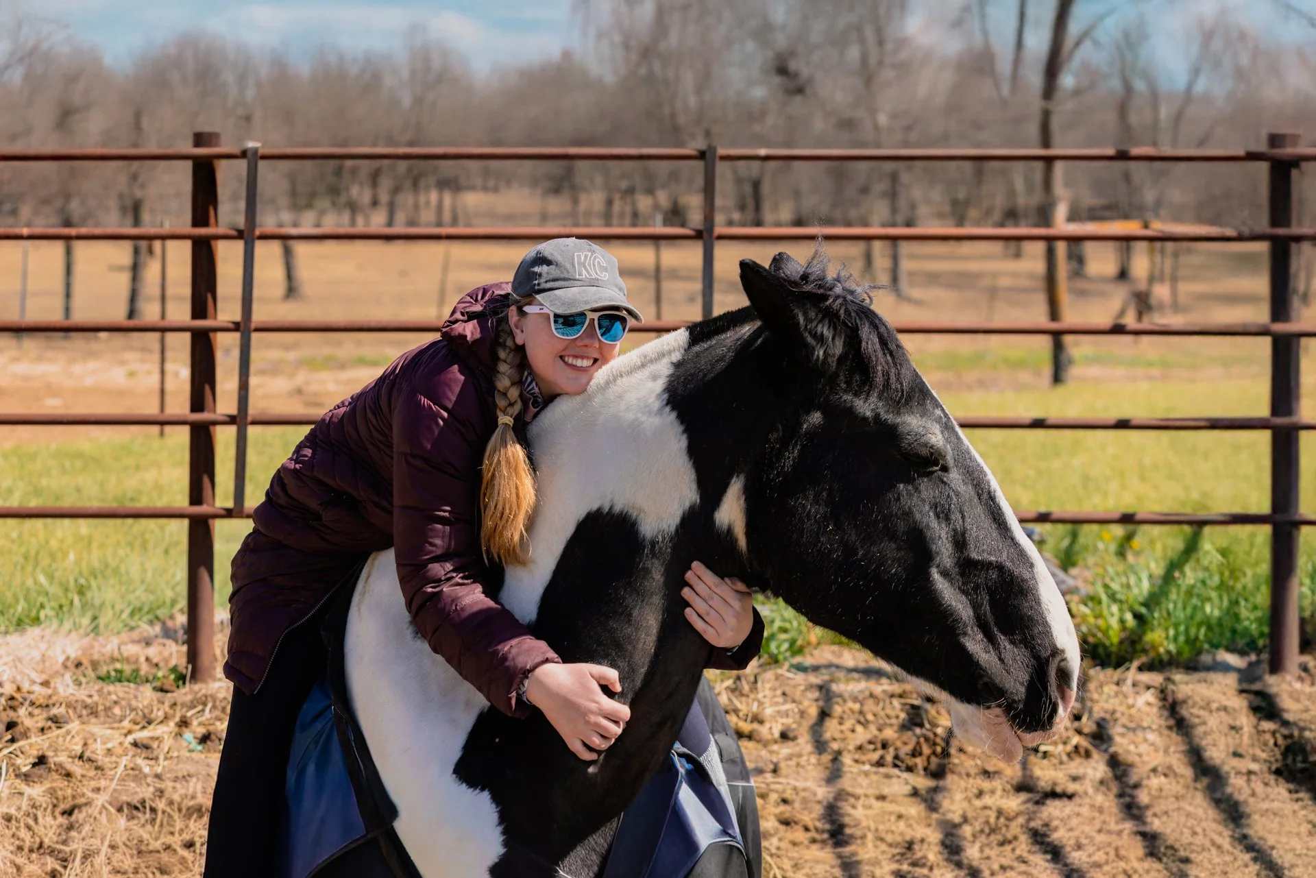 A woman with blonde hair in braids, wearing sunglasses, a gray baseball cap, and a maroon jacket, hugging a black and white horse in an outdoor paddock.