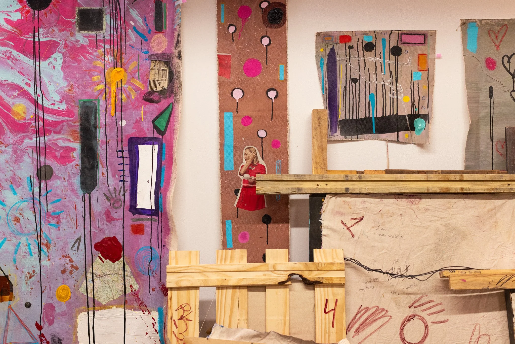 Mixed media art installation with colorful abstract paintings on the wall, including pink, black, cyan, and purple elements, with a small cutout photograph of a woman in a red dress. Various wooden and cardboard materials are arranged in front of the