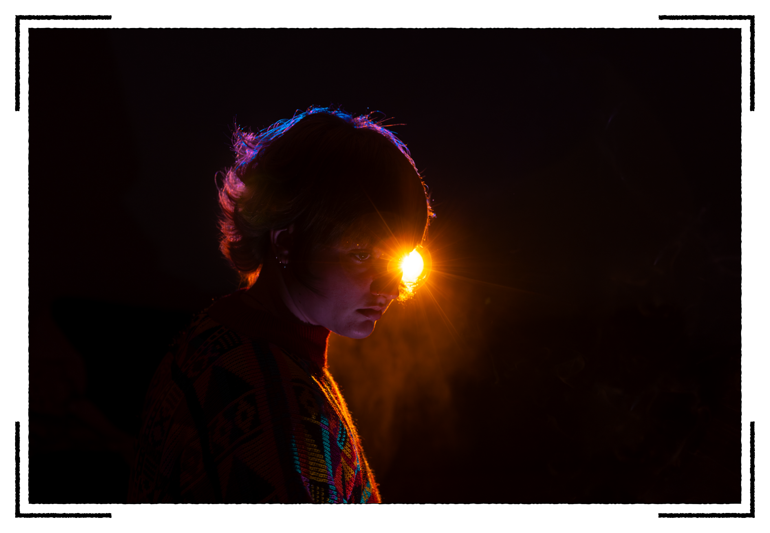 A person with short hair and colorful sweater is looking to the side in a dark background, with a bright light shining on their face from their right eye resulting in a starburst effect.