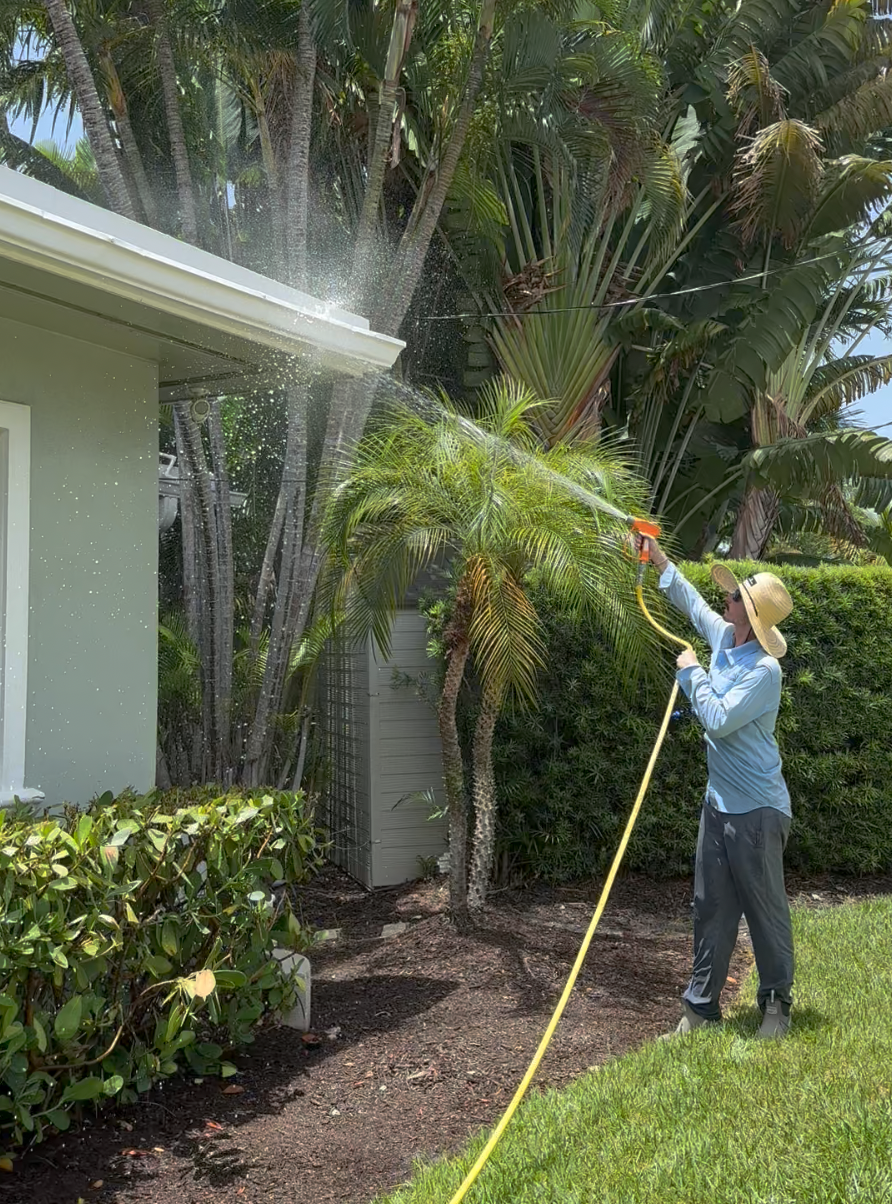Person watering plants outside a house with a garden hose, wearing a wide-brimmed hat, long-sleeve shirt, and pants, surrounded by greenery and trees.