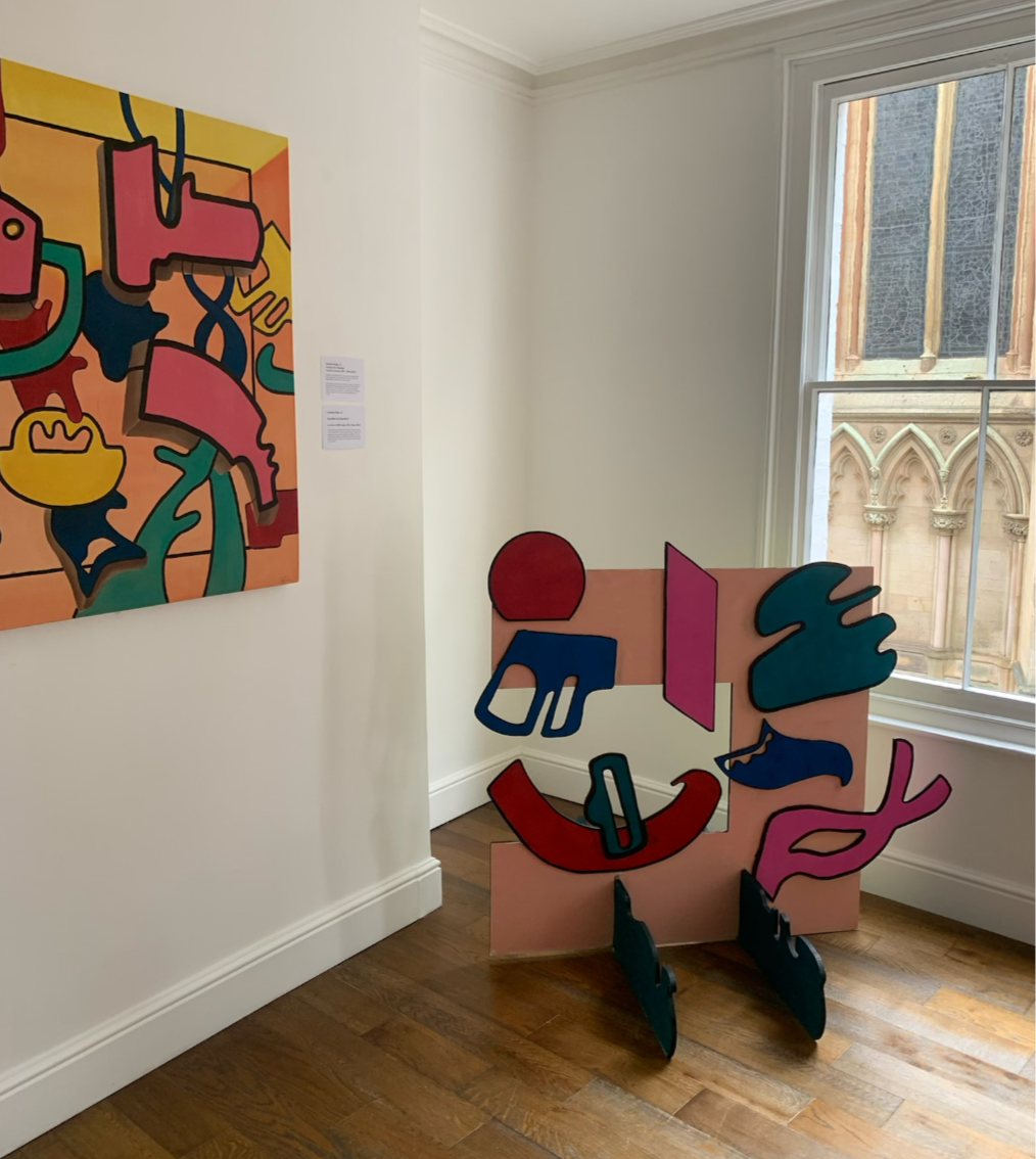 An art gallery scene with a colorful abstract painting on the left wall and a sculptural installation on the floor near a window. The sculpture features various abstract shapes in pink, blue, red, and black mounted on a pink base. A large window reveals an exterior view of a historic building with ornate stone architecture.