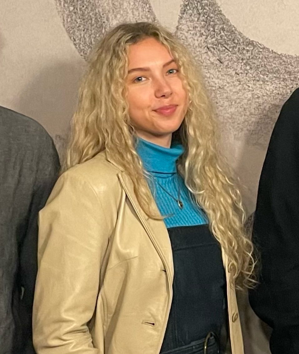 A woman with curly blonde hair smiling, wearing a beige leather jacket over a blue turtleneck, Gabriela Stelpe