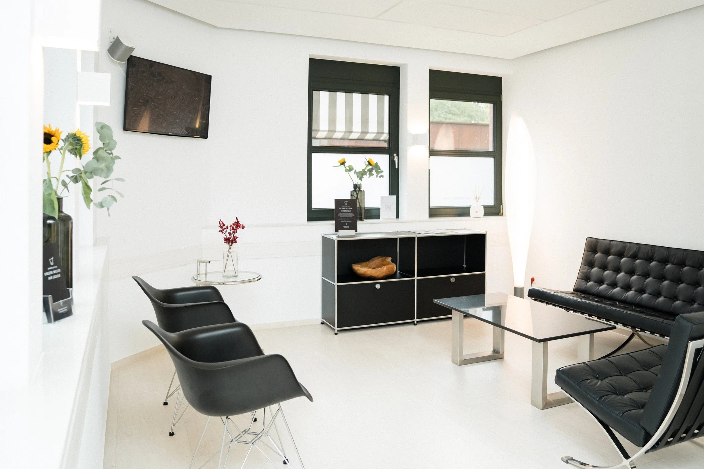 Ein modernes, helles Warteraum mit schwarzem Ledersofa, mehreren schwarzen Stühlen, einer schwarzen Kommode mit Dekorationsgegenständen und Sonnenblumen in einer Vase vor einem Fenster.