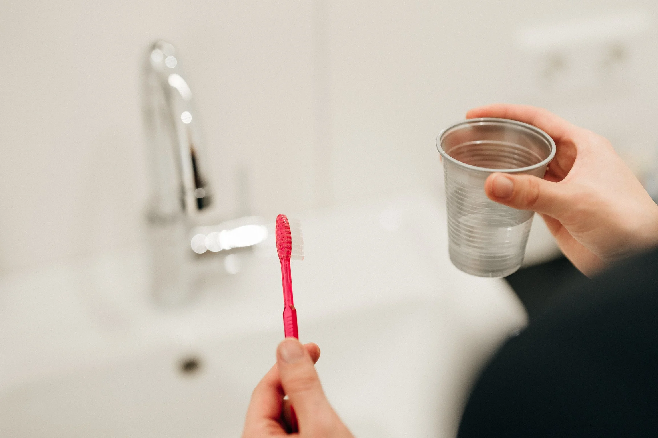 Die Hand einer Person hält einen leeren Metallbecher, während eine andere Hand einen rosa Zahnbürstenkopf hält. Der Hintergrund zeigt eine weiße Wand mit einem Wasserhahn.