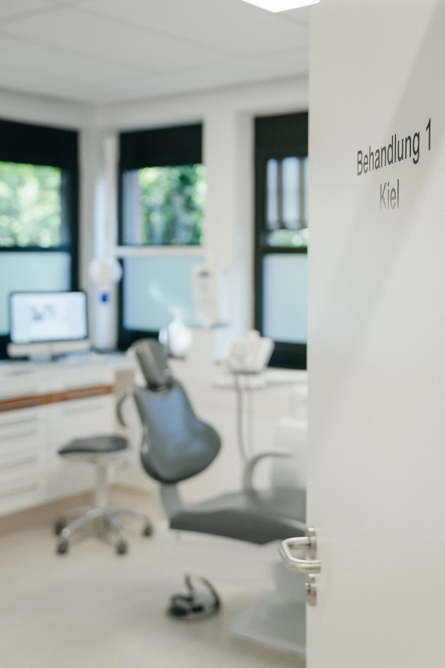 Ein Behandlungszimmer im zahnmedizinischen Bereich mit einem weißen Schreibtisch, einem weißen Stuhl, einem Behandlungsstuhl und einem Monitor. An der Tür hängt ein Schild mit der Aufschrift 'Behandlung 1 Kiel'.