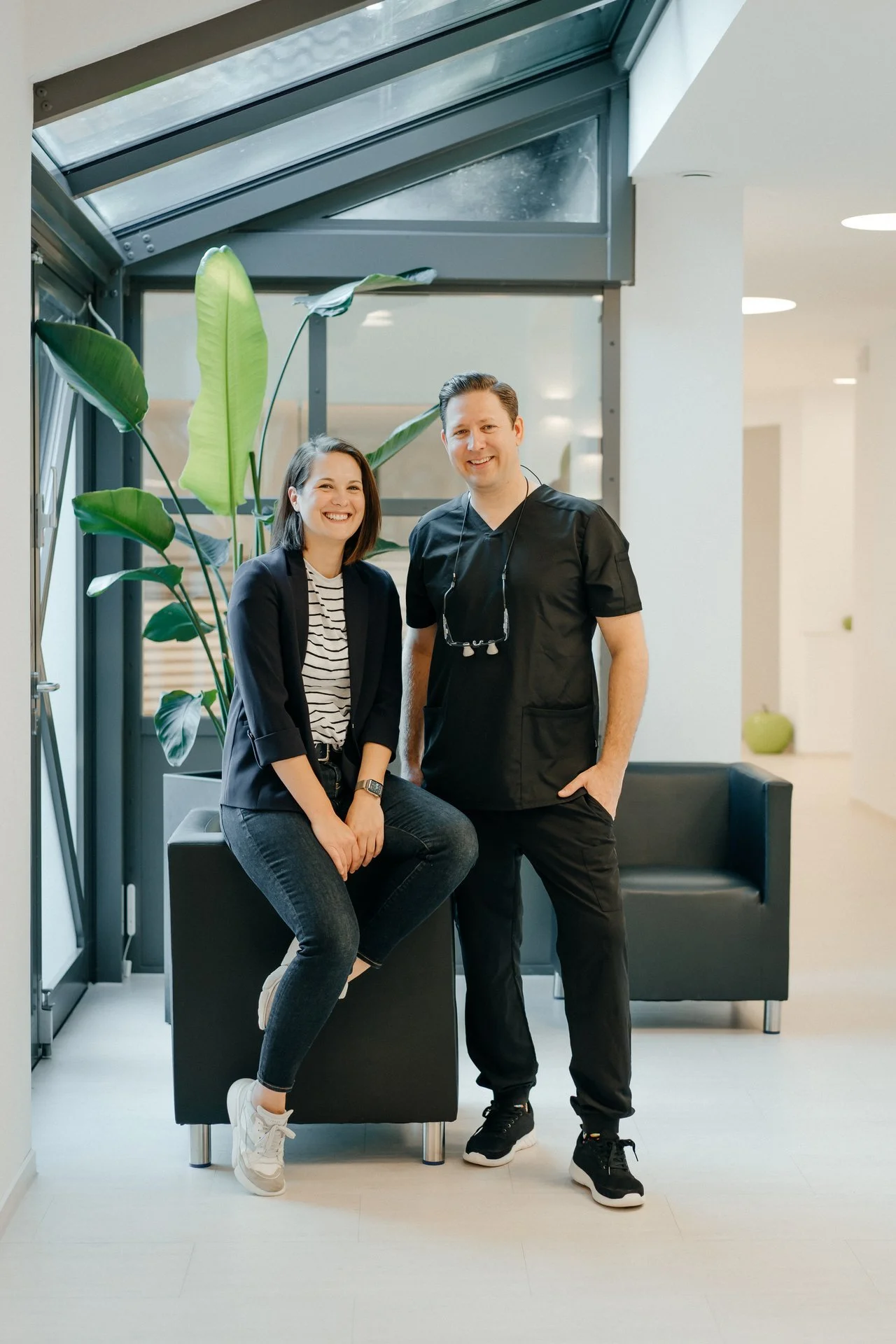 Zahnarzt Dany Zoubie und Praxismanagerin Anja Zoubie, beide lächeln. Im Hintergrund große grüne Pflanzen und moderne Einrichtung in einem hellen Raum.