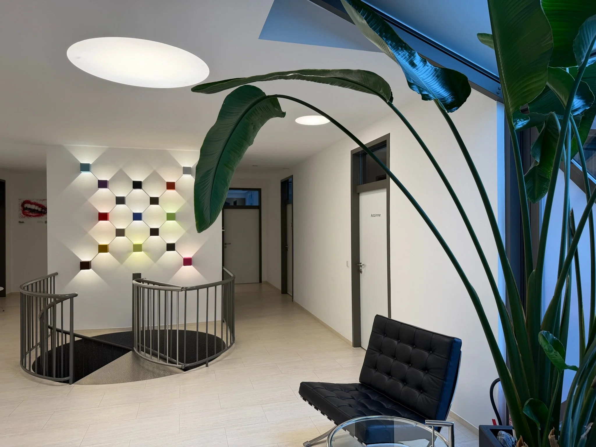 Ein heller Raum mit weißen Wänden und einer Treppe im Vordergrund. An der Wand sind geometrische Wandlampen in verschiedenen Farben angebracht. Es gibt große grüne Pflanzen und moderne schwarze Sitzmöbel.