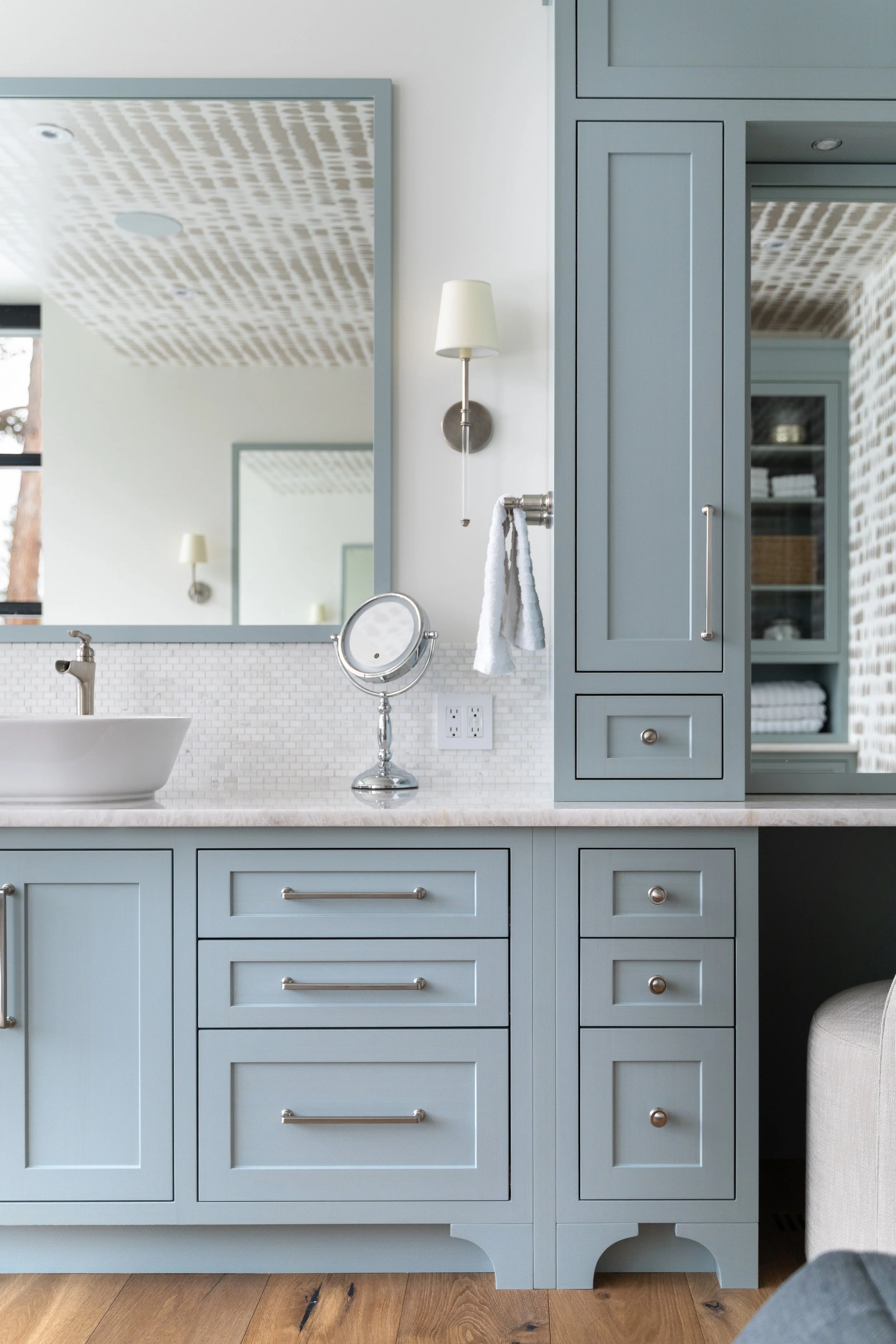 A bathroom vanity with light blue cabinetry, a white vessel sink, a small round mirror, and a wall-mounted light fixture with a white shade. There is a large mirror above the sink, and a section of open shelving with towels and baskets. The lighting 