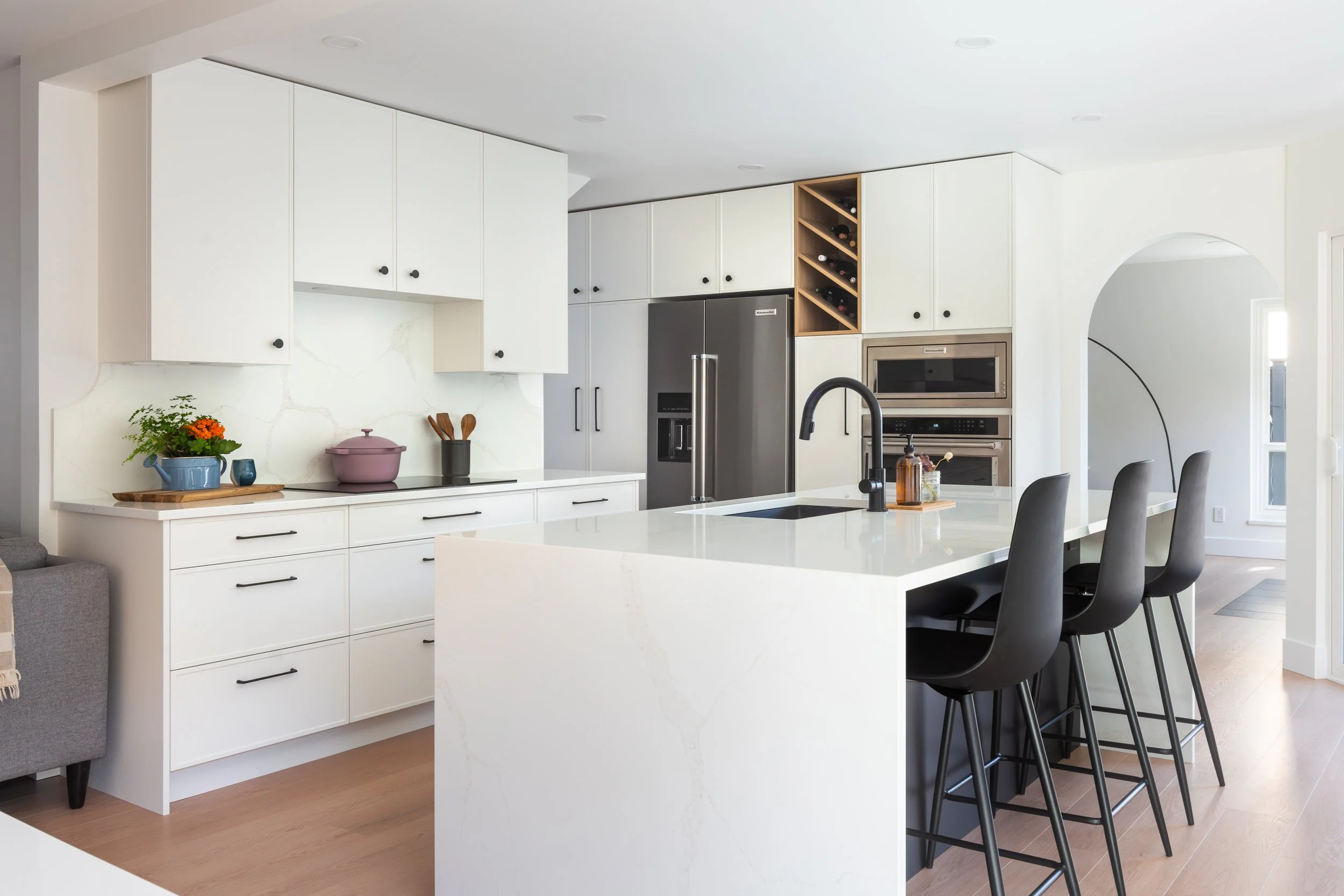Modern white kitchen with black barstools, stainless steel refrigerator, built-in oven, and marble island.