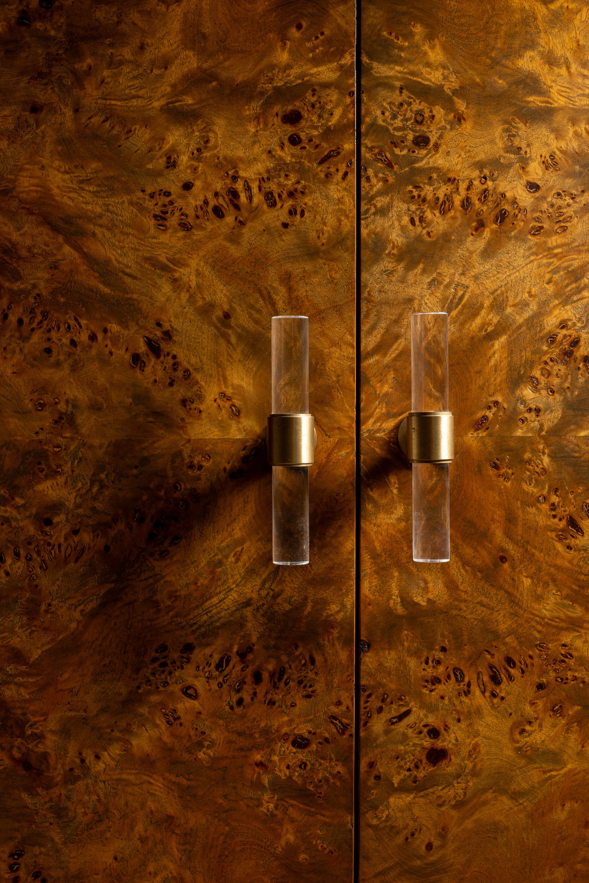Close-up of a wooden cabinet with a burl wood pattern, featuring two vertical glass and brass handles.