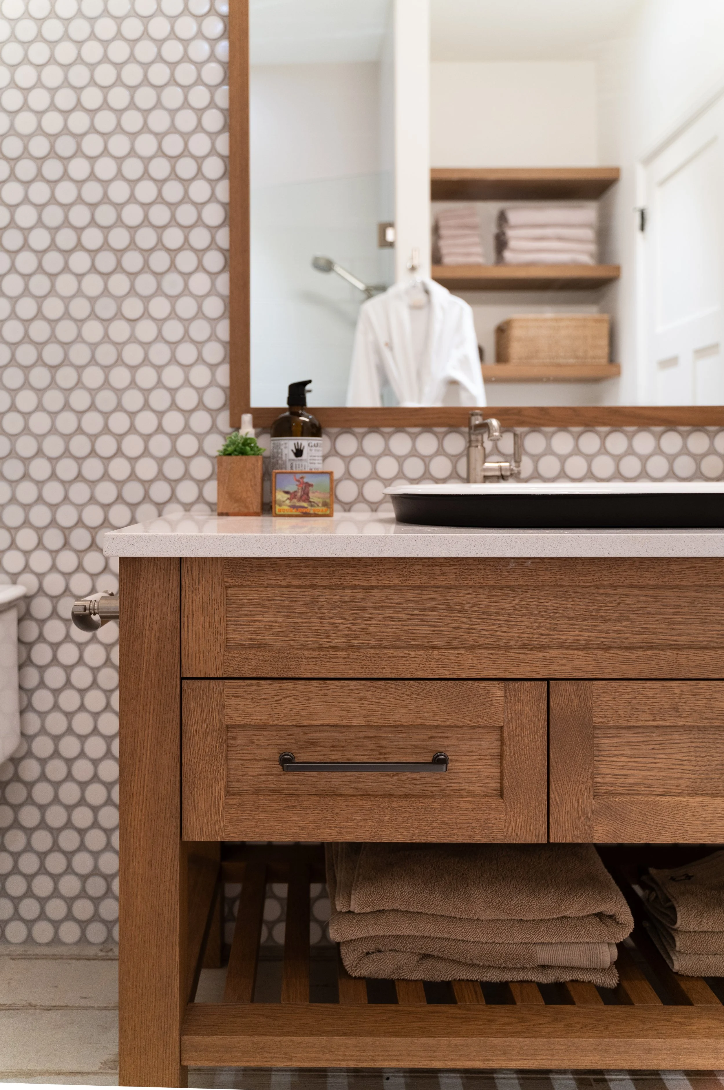 Bathroom vanity with a mirror, wooden cabinet, countertop with a soap dispenser, small plant, and a note or small framed picture, with shelves holding towels and other items in the background.