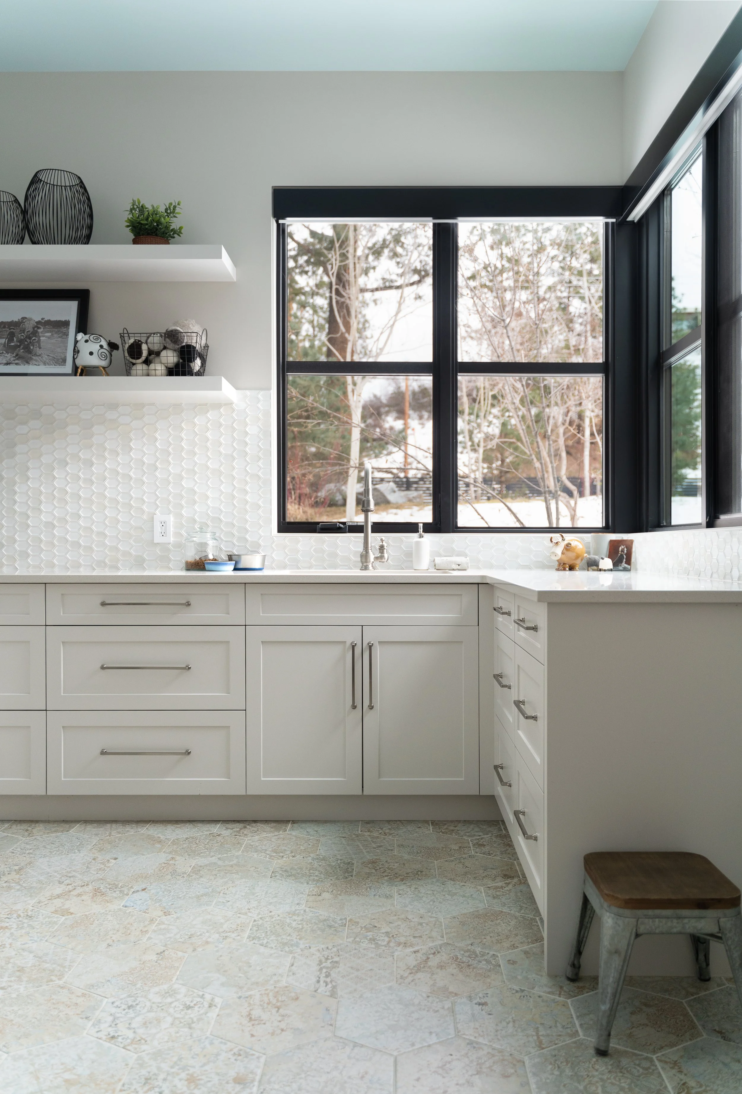 Kitchen with white cabinets, a large window, and a tiled backsplash. Decor includes black wire vases, a small plant, framed photo, and kitchen essentials on the countertop. Natural light through the window illuminates the space.