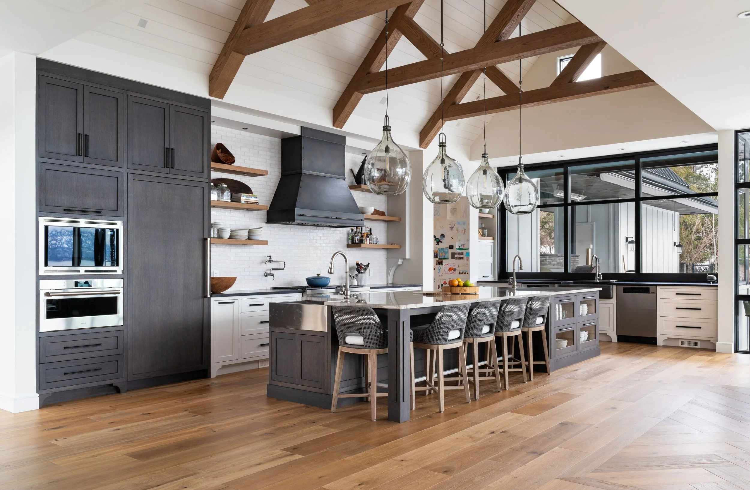 Modern kitchen with wooden beams, dark cabinets, and a large island with barstools, overlooking a bright space with large windows and hardwood floors.