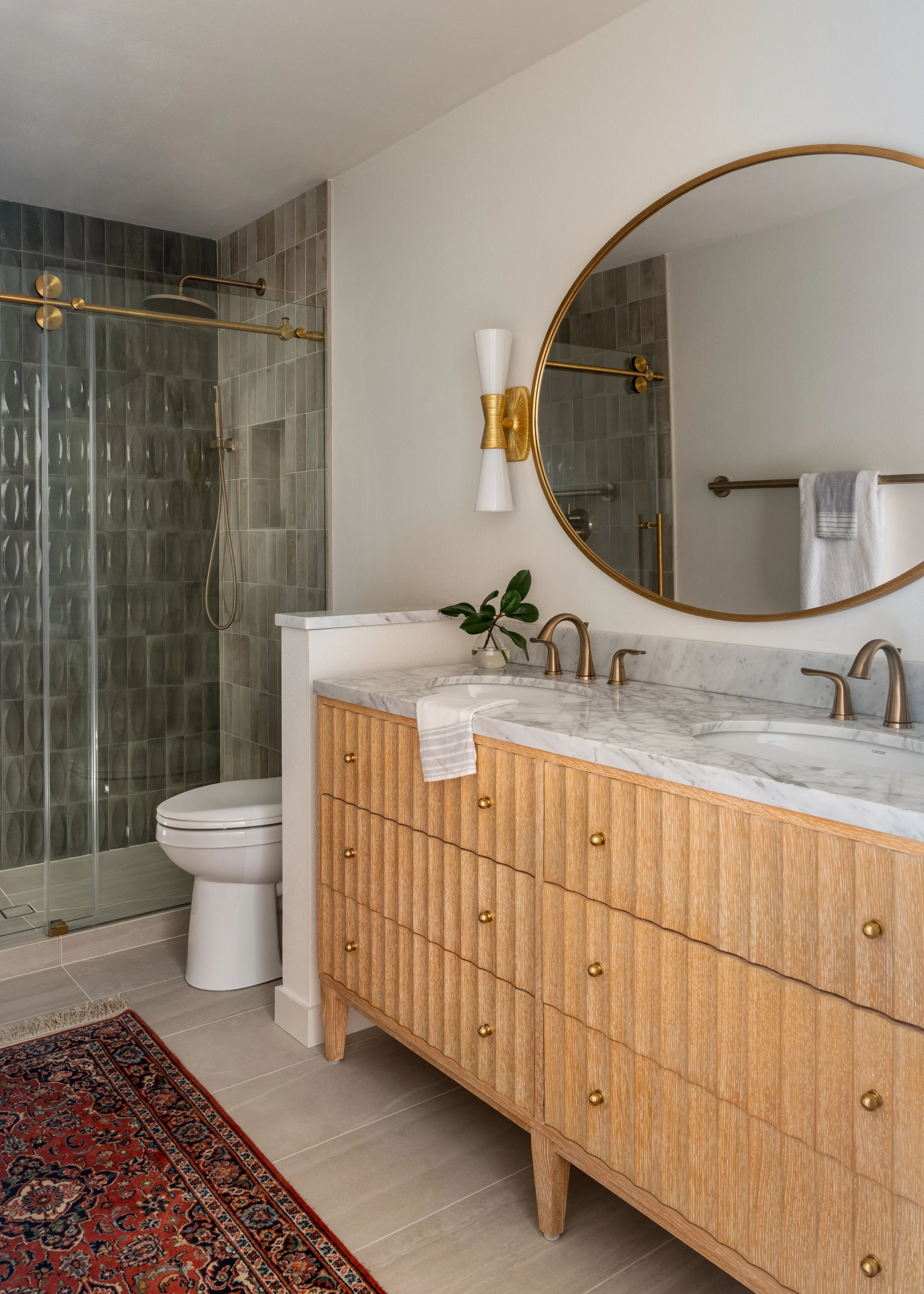 Primary bathroom on Bainbridge Island featuring a fluted wood vanity, marble double sinks, brass fixtures, and textured green tiles.