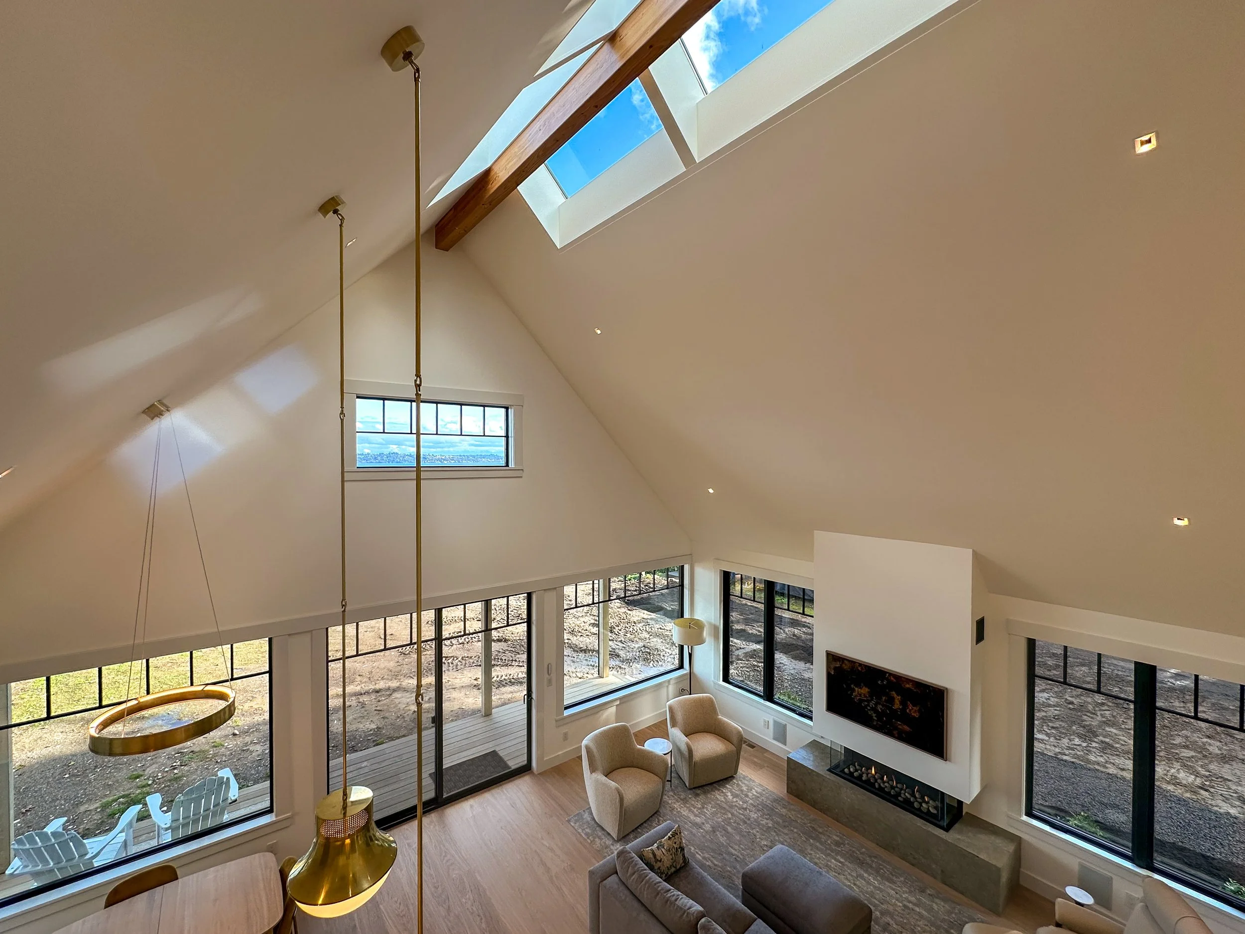 Photo of a modern living room with high vaulted ceiling, large windows, and a skylight. The room has beige chairs, a sofa, a fireplace, and a wood floor. There are outdoor chairs visible through the windows.