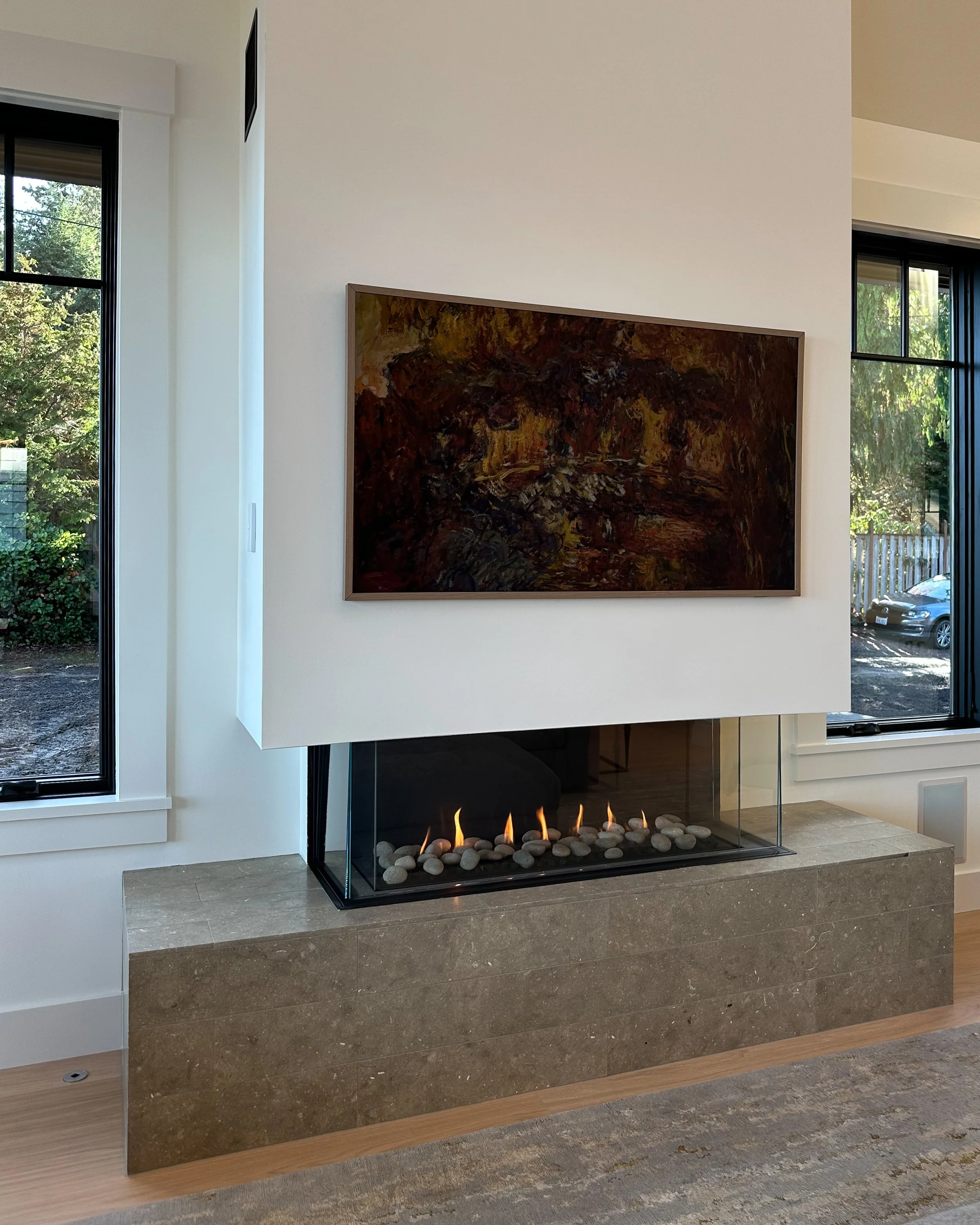 Modern living room fireplace with a framed abstract painting above a glass-enclosed gas fire with rocks, set on a stone surround, next to large windows showing greenery outside.