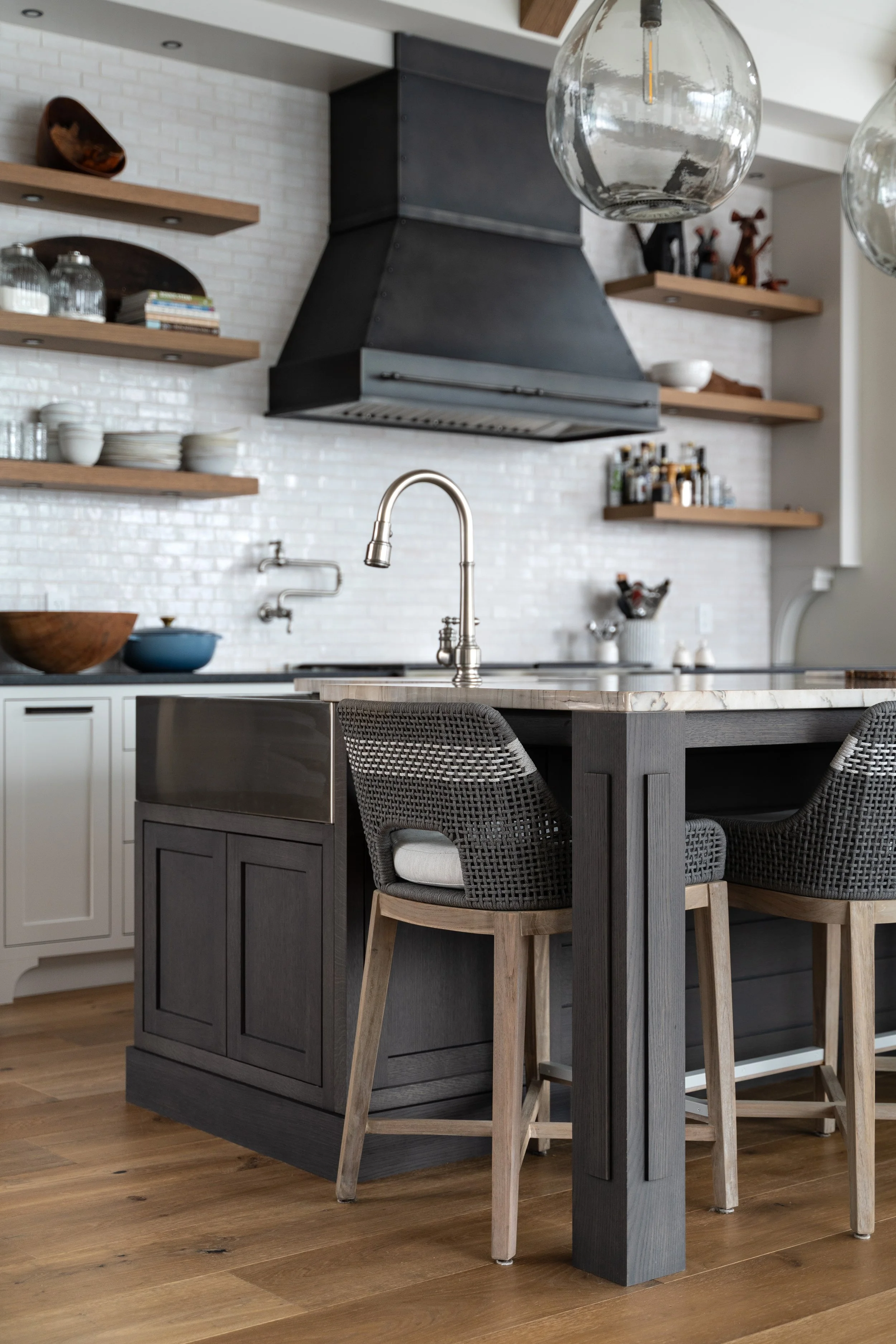 Modern kitchen with white brick backsplash, black range hood, open wooden shelving with glassware, dishes, and bottles; center island with marble countertop, black base, stainless steel sink, and barstools with woven backs and wooden legs; pendant li