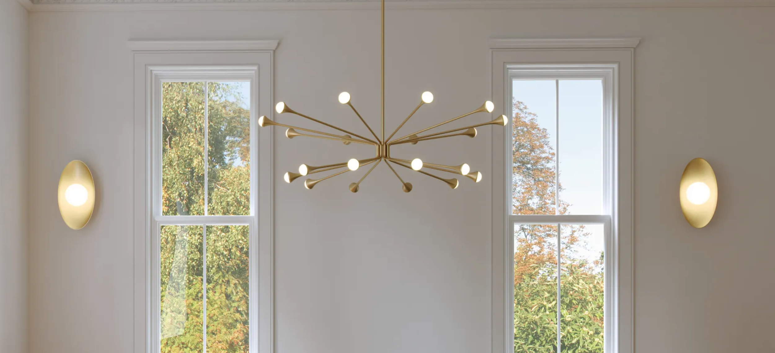 Interior room with white walls, two large windows, a modern gold chandelier with multiple arms and lights, and wall-mounted sconces on either side of the windows.