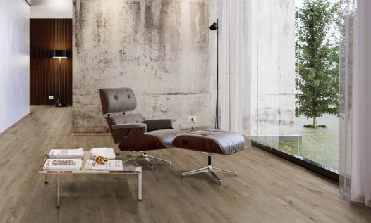 Modern interior room with a gray lounge chair and matching ottoman, a small white table with newspapers, and large windows offering a view of trees outside.