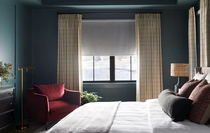 Bedroom with large window, beige curtains, a burgundy armchair, a white bed with various pillows, and a nightstand with a lamp.