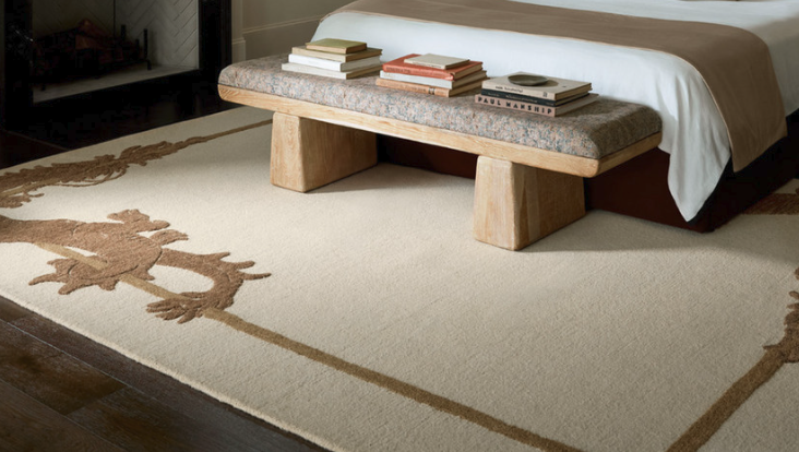 A bedroom with a large beige rug, a bed with a bench at the foot, and a stack of books on the bench.