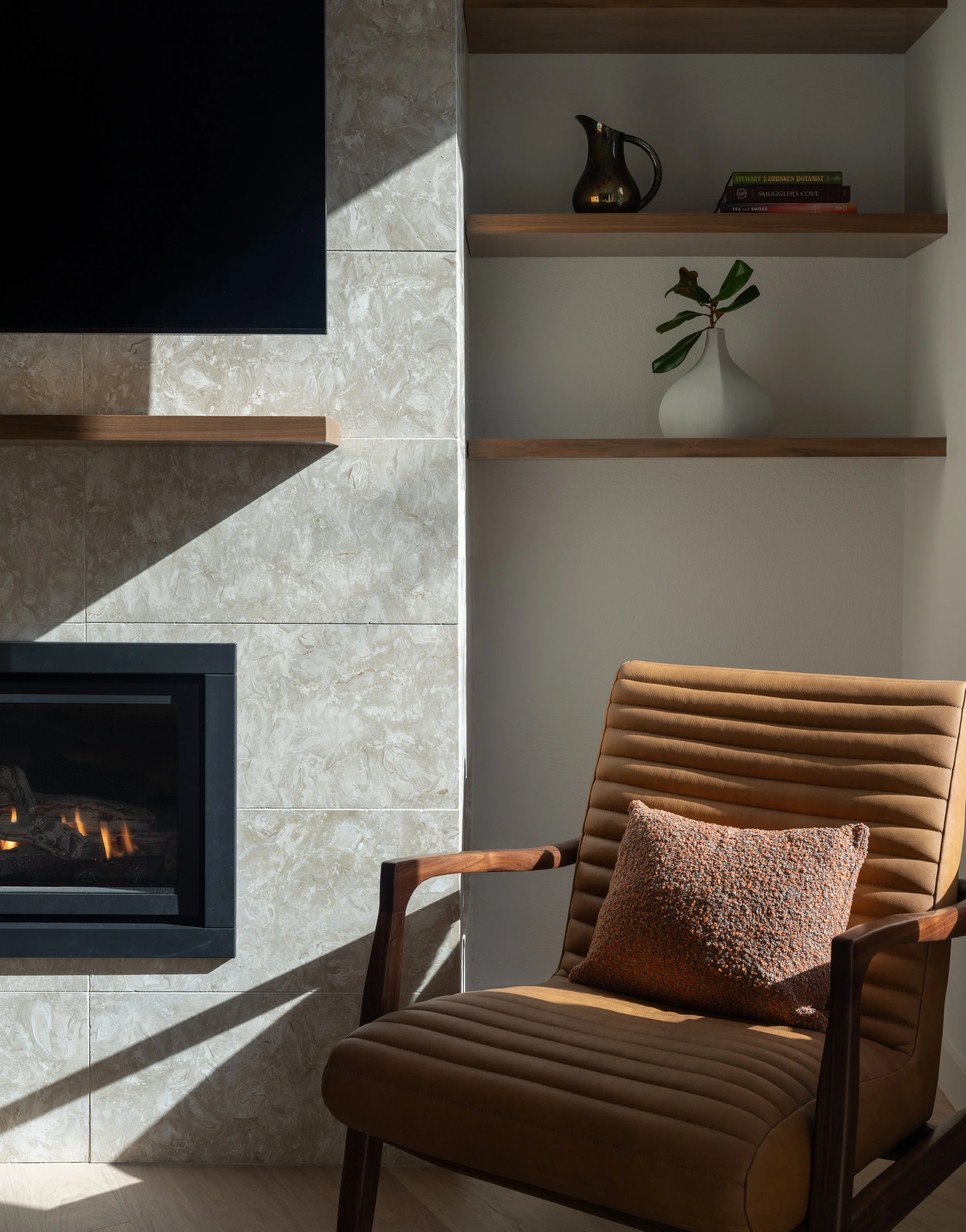 Modern gas fireplace with a textured stone tile surround, floating wood shelves, and a tan mid-century leather armchair on Bainbridge Island.
