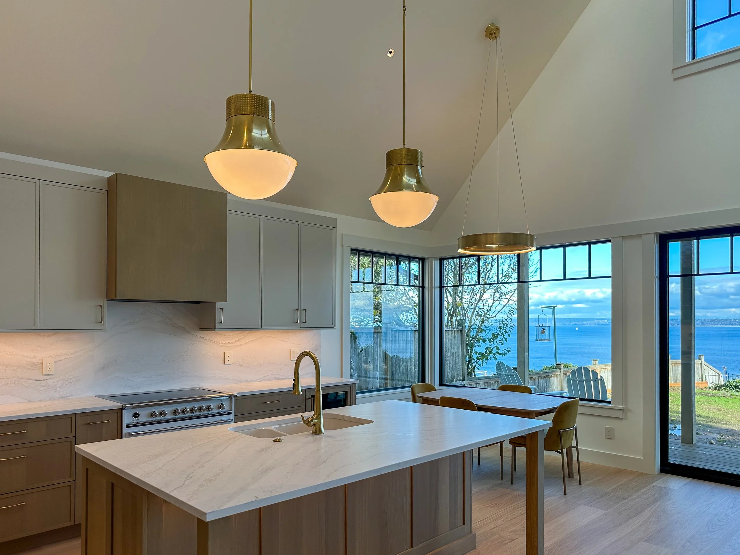 Modern kitchen with island, brass pendant lights, and large windows overlooking a lake, with a small dining table and chairs.
