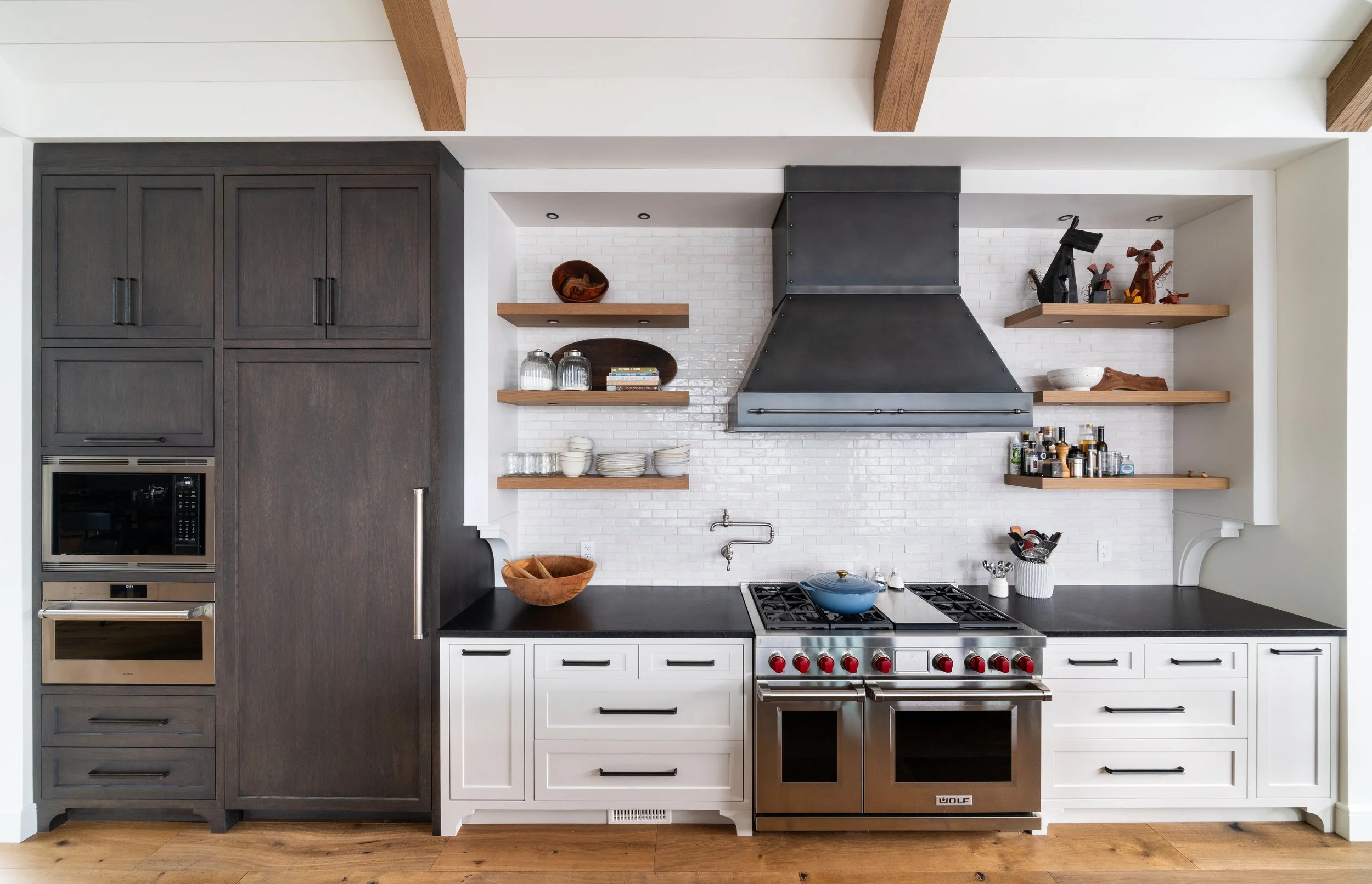 Modern kitchen with dark wood cabinetry, white shiplap wall, black countertop, and open wooden shelves displaying dishes, glassware, and decorative items. A stainless steel Wolf stove with red knobs is centered under a black metal range hood.