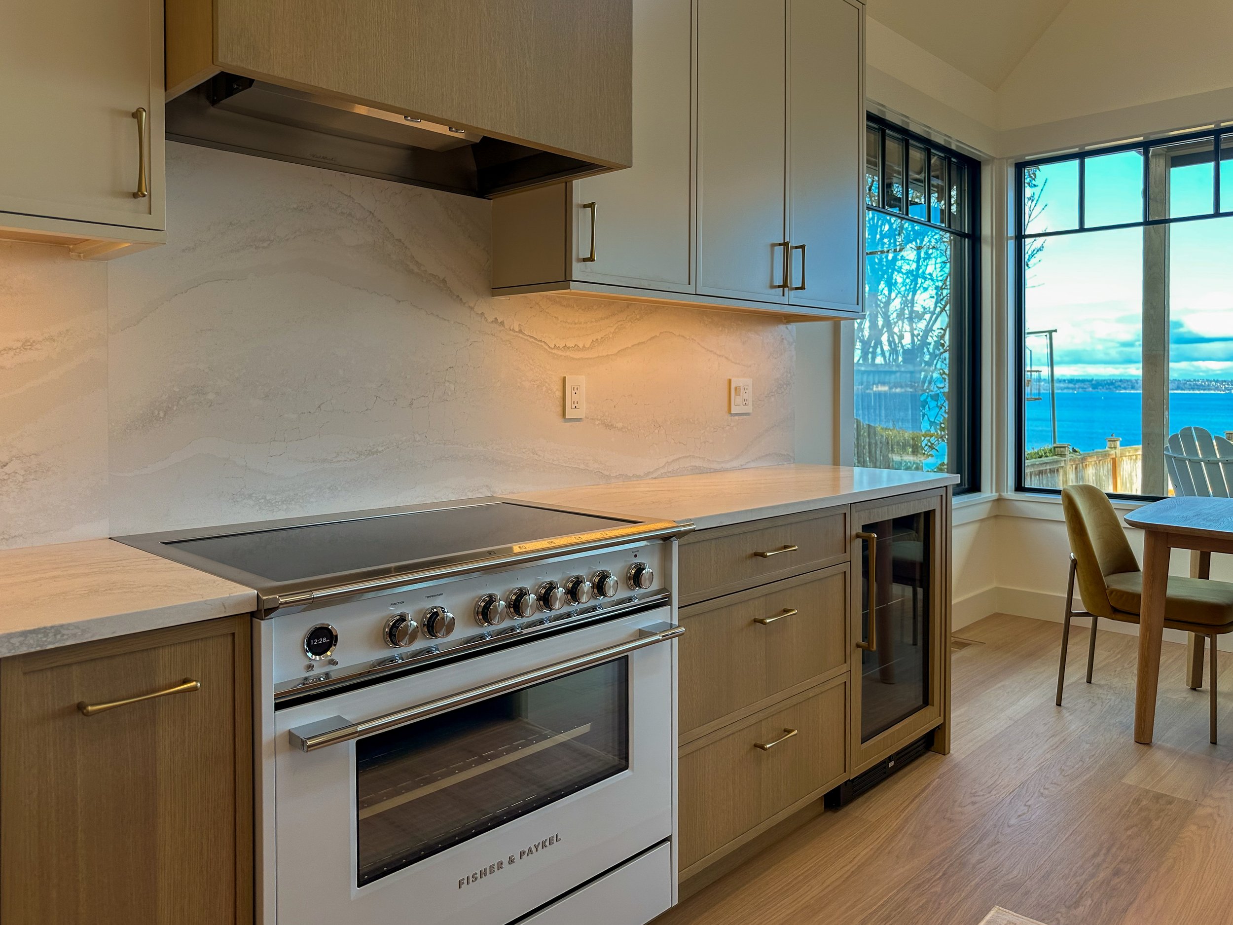 Kitchen with wooden cabinets, a white oven, marble countertops, large windows showing a view of water and sky, yellow chairs around a wooden table.