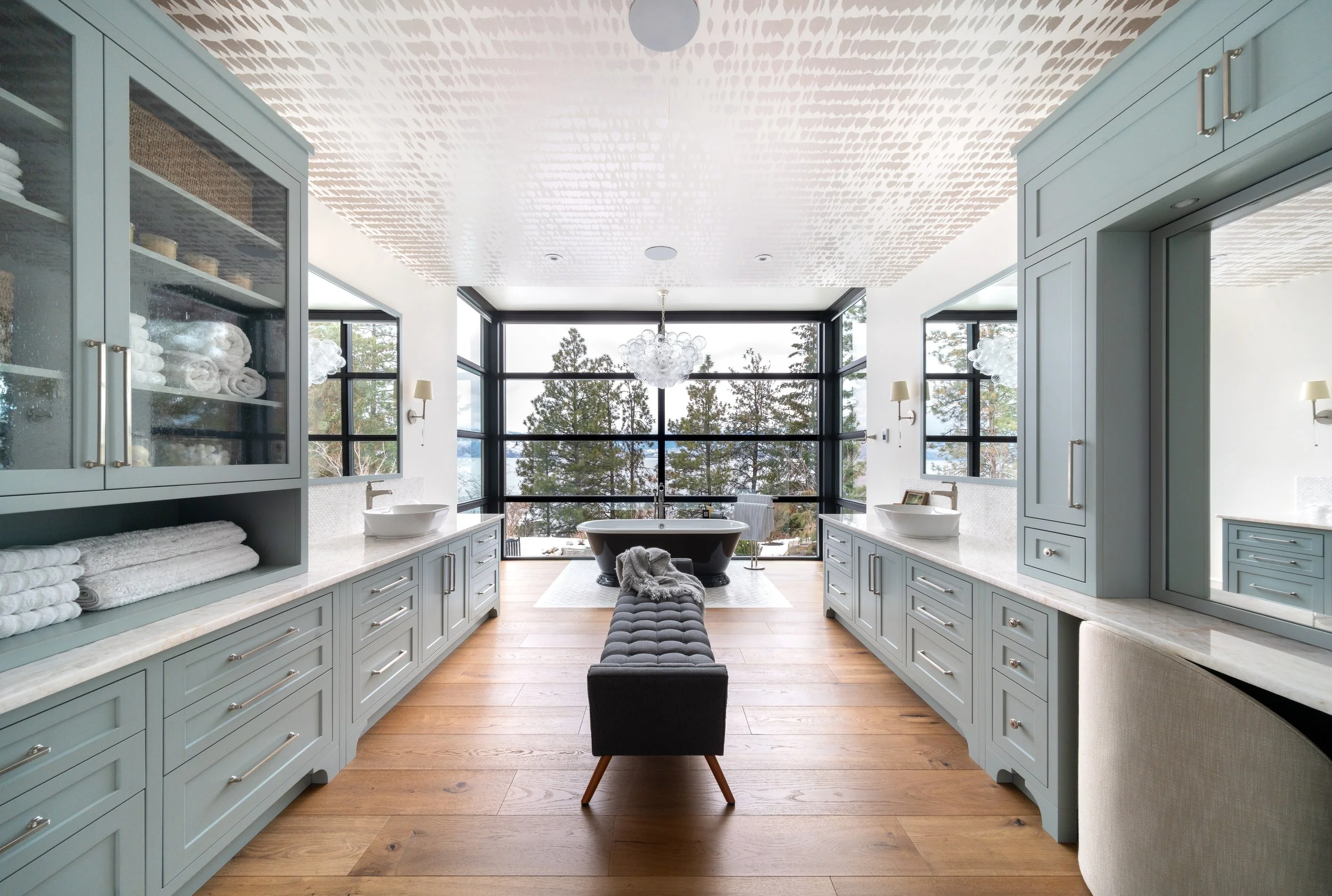 Modern bathroom with light blue cabinetry, large windows, a bathtub, and wooden floors.