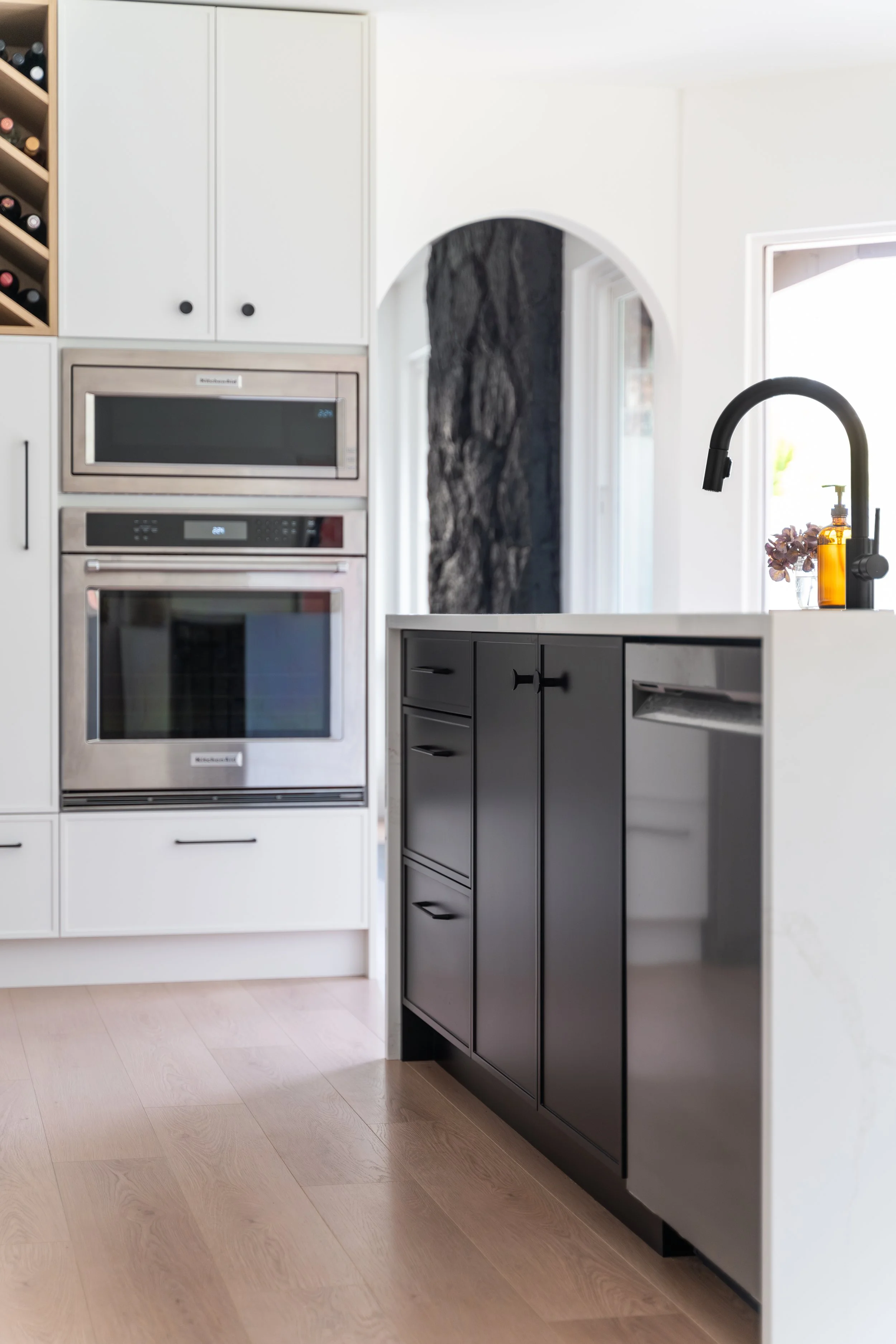 Modern kitchen with black and white cabinetry, built-in stainless steel oven and microwave, black faucet, white countertops, a small brown bottle with pump, and a window in the background.