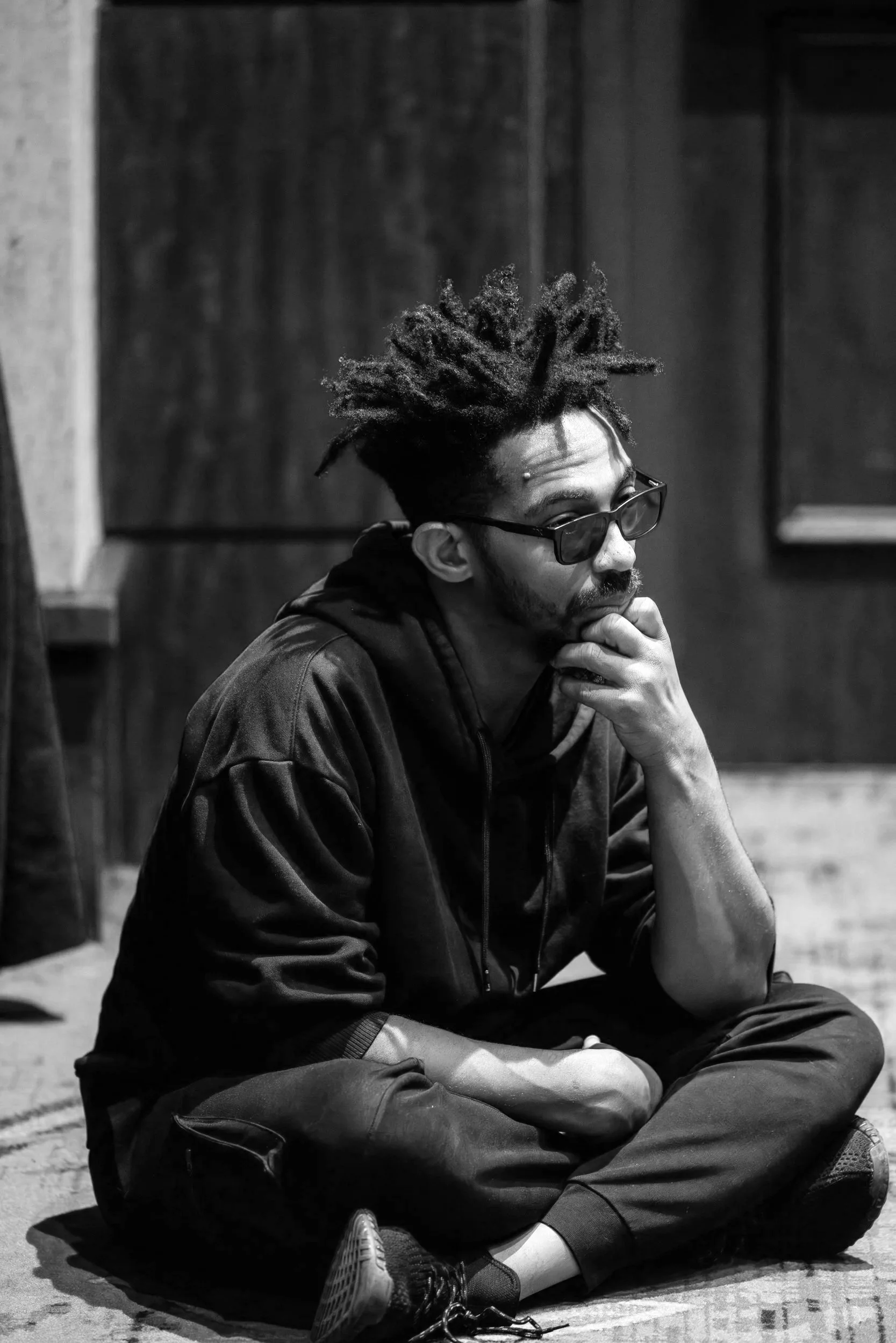 A young man with dreadlocks, wearing glasses, a jacket, and sweatpants, sitting on the ground in a contemplative pose with his hand resting on his chin indoors, black and white photo