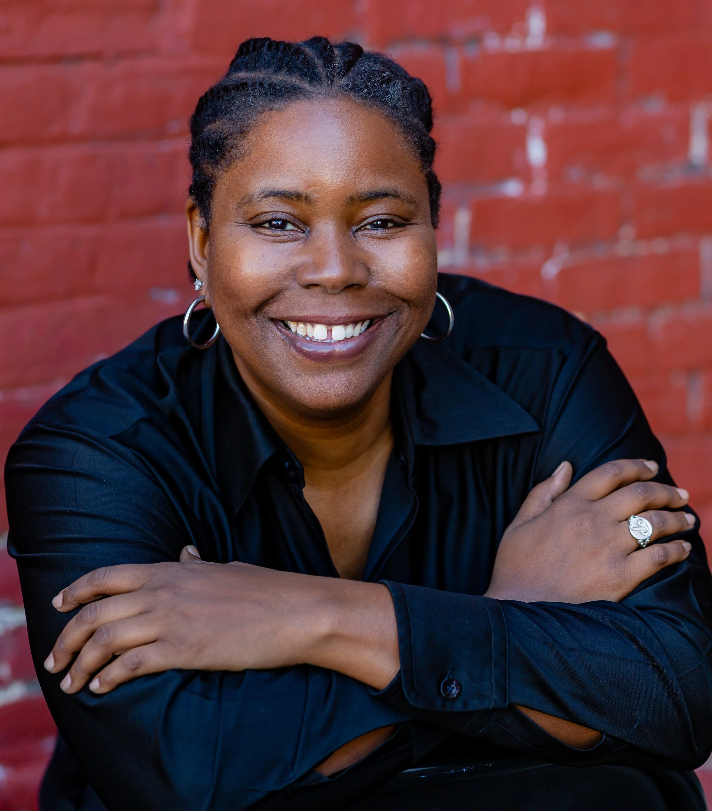 Close-up of a smiling African American woman with braided hair, wearing hoop earrings, a silver ring, and a black shirt, standing against a brick wall.