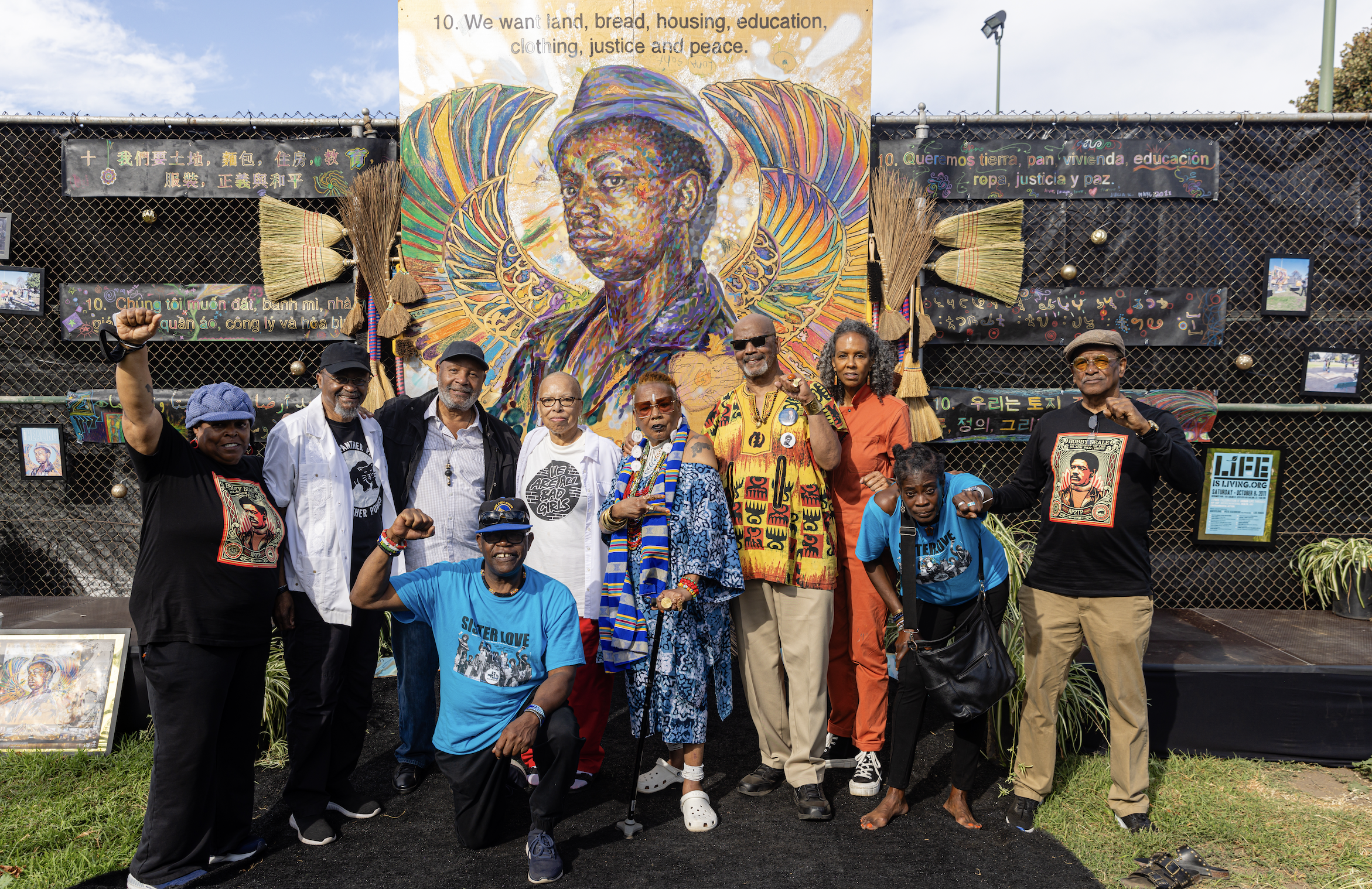 A group of diverse people standing in front of a large colorful mural of a woman with artwork and banners around them, some raising fists, at a community or activist event.