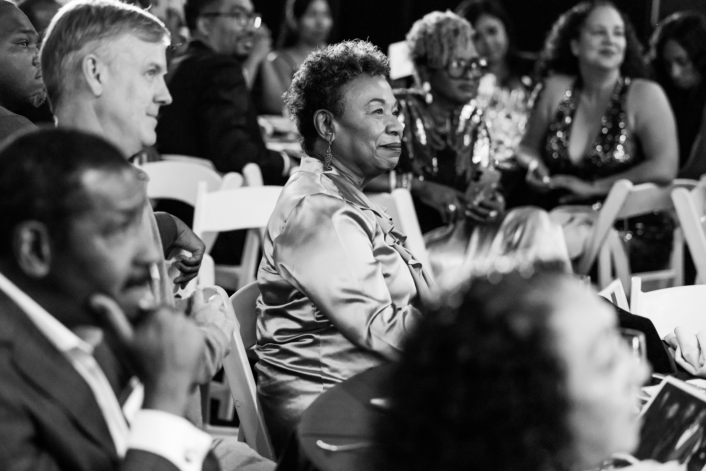 A group of diverse adults sitting in an audience, attentively listening at an event. Focus on an older woman with short, curly hair and earrings, seated in the center, smiling softly.
