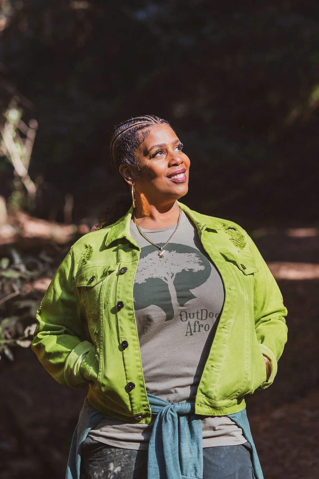 A woman outdoors in sunlight, wearing a bright green jacket over a t-shirt that reads 'OutDog Afro', with a denim shirt tied around her waist, looking up with a smile.