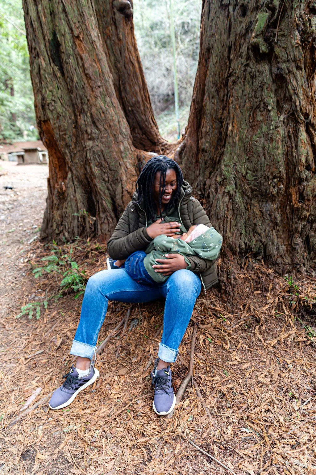 A woman in jeans, sneakers, and a jacket sits on the forest ground, holding and smiling at a baby wrapped in a green blanket, with a large tree trunk behind them.