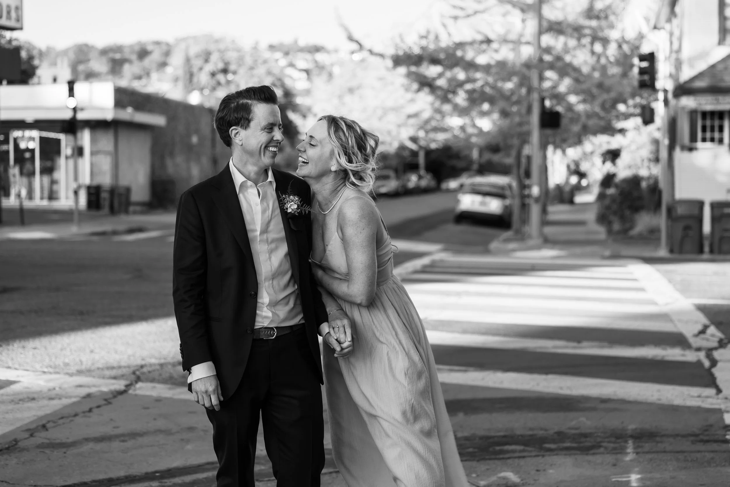 A black-and-white photo of a couple smiling and holding hands while walking across a crosswalk on a city street.