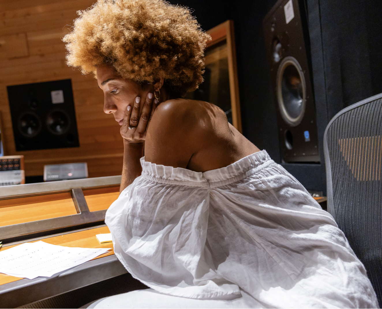 A woman with curly, blonde hair sitting at a music studio desk, resting her chin on her hand, surrounded by audio equipment and a wooden wall.