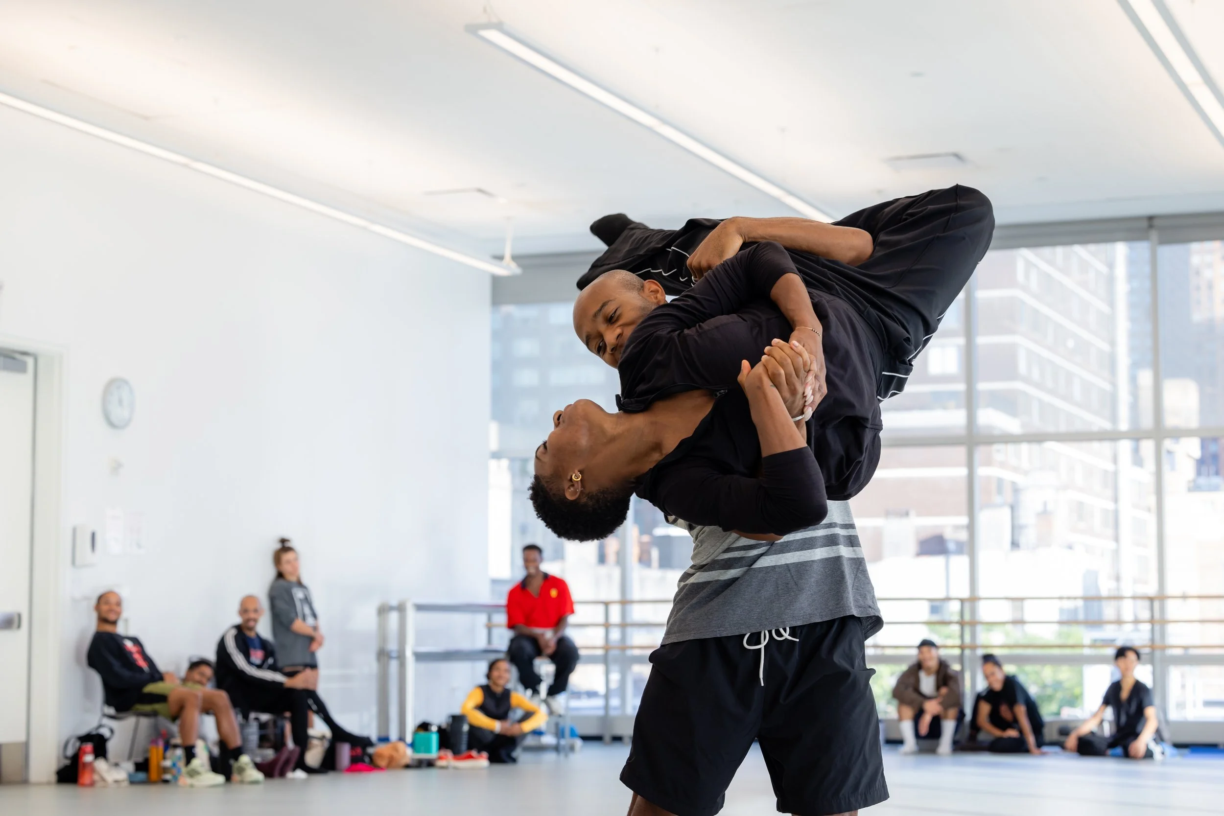 Two men practicing Brazilian Jiu-Jitsu in a martial arts class, one holding the other in a ground position while a group of people watches in a spacious, bright studio with large windows.