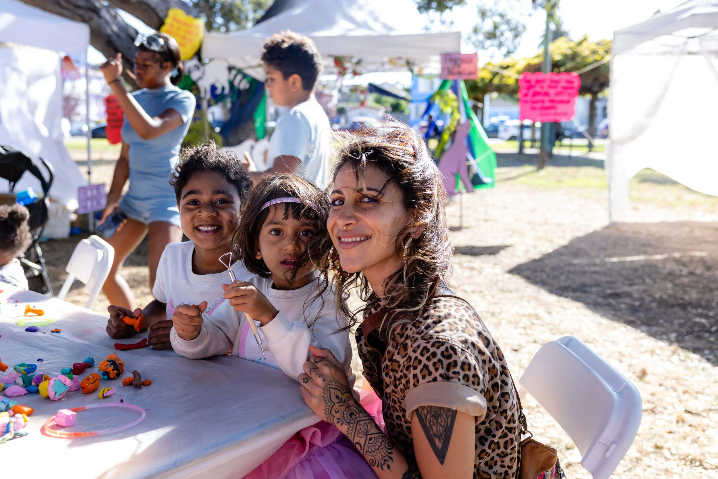 A woman with tattoos and leopard print clothing sitting at a table with three children, all smiling, at an outdoor event with tents and colorful signs in the background.