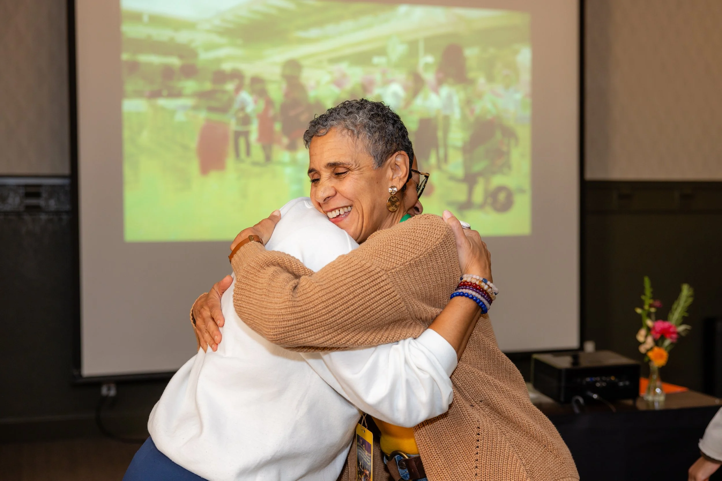 Two women are hugging each other and smiling in a room with a blurred background that includes a projection screen and a vase of flowers on a table.