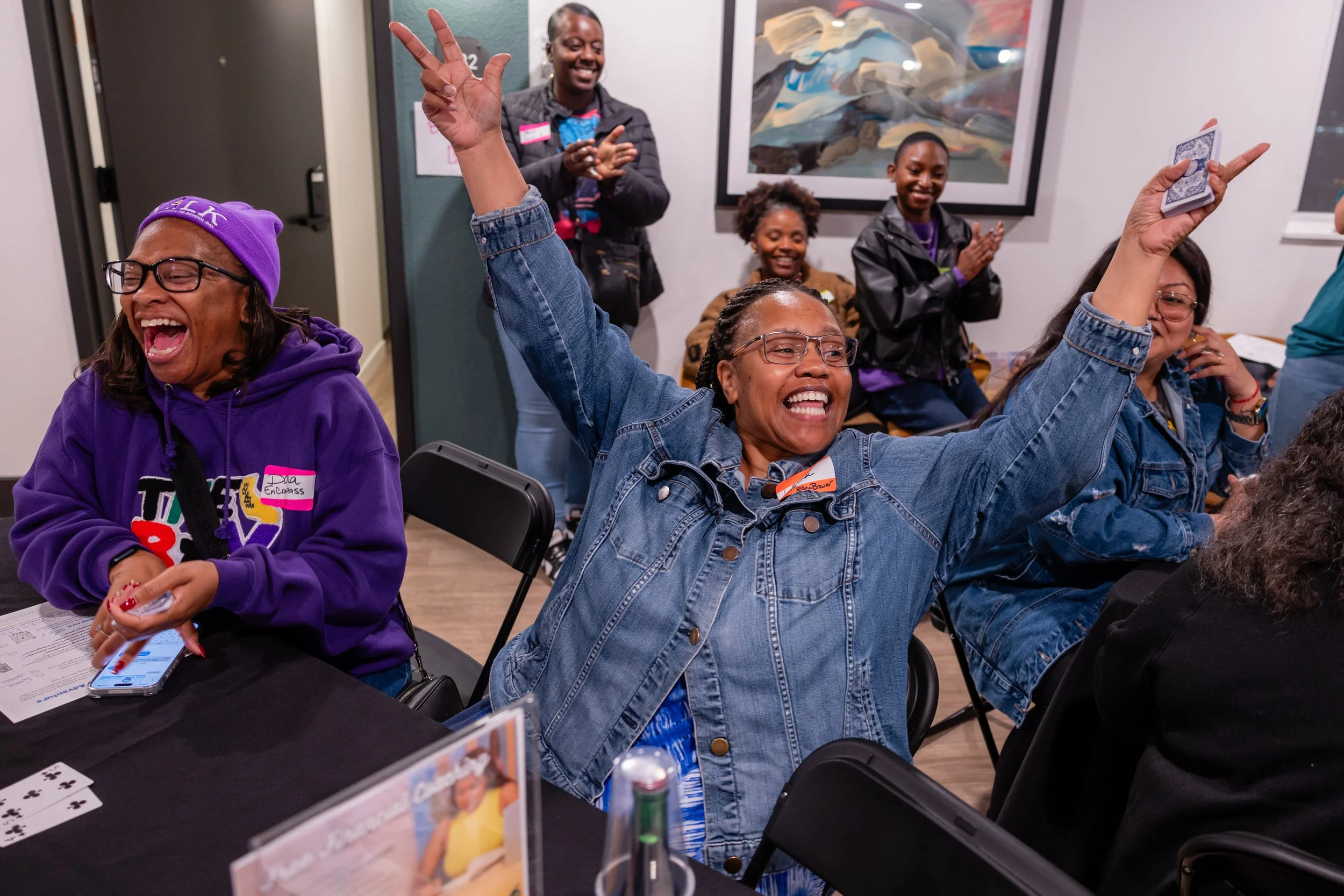 A group of women enjoying a lively moment at a social gathering, with one woman raising her arm and smiling widely, while others claps and laugh in the background. The setting appears to be indoors, possibly a community center or event space, with ar