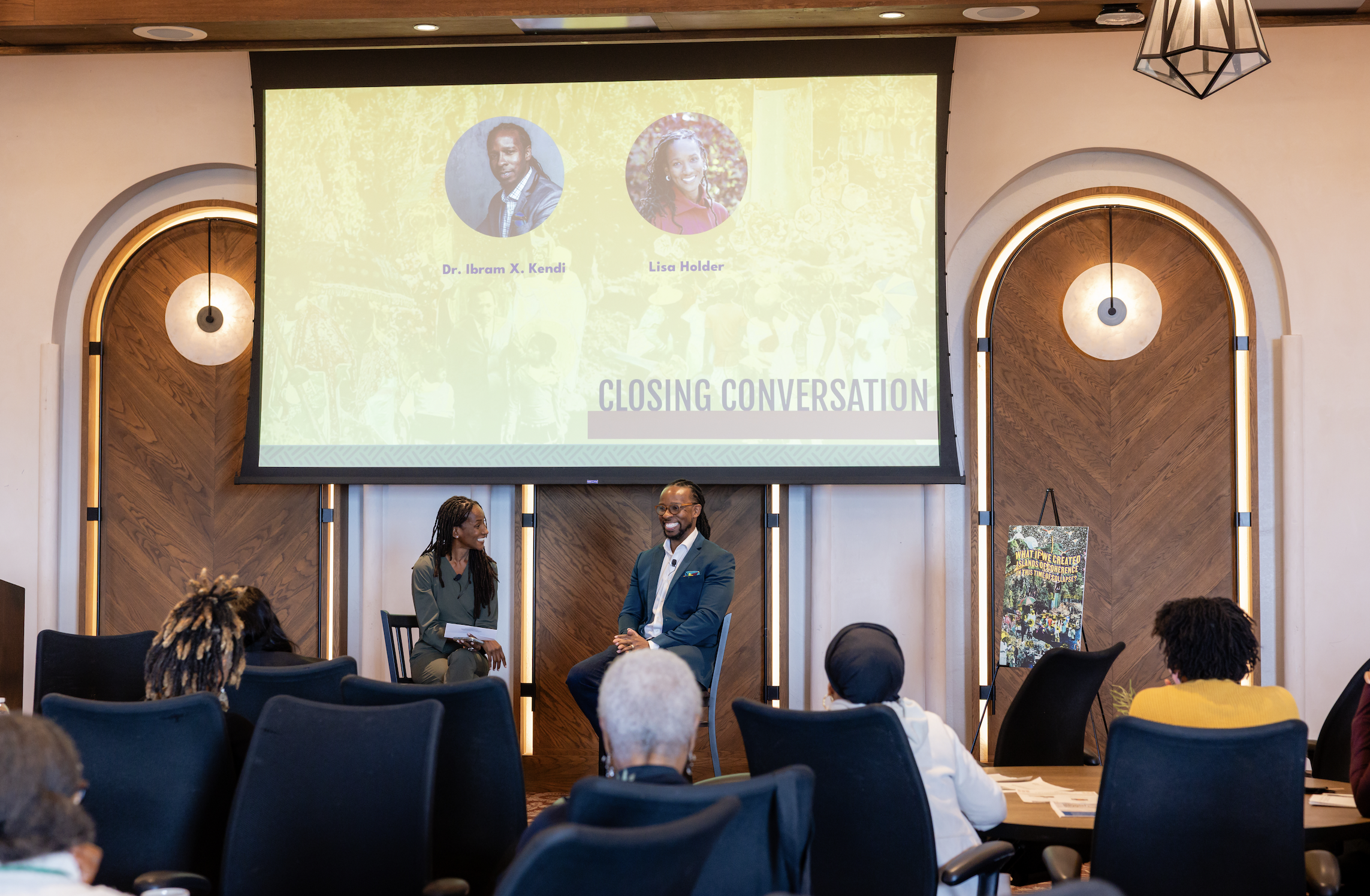 A conference room with a large screen displaying two photographs and names of speakers, Dr. Ibram X. Kendi and Lisa Holder, during a closing conversation. The room has wood paneling, round wall lights, and audience members seated facing the stage, in