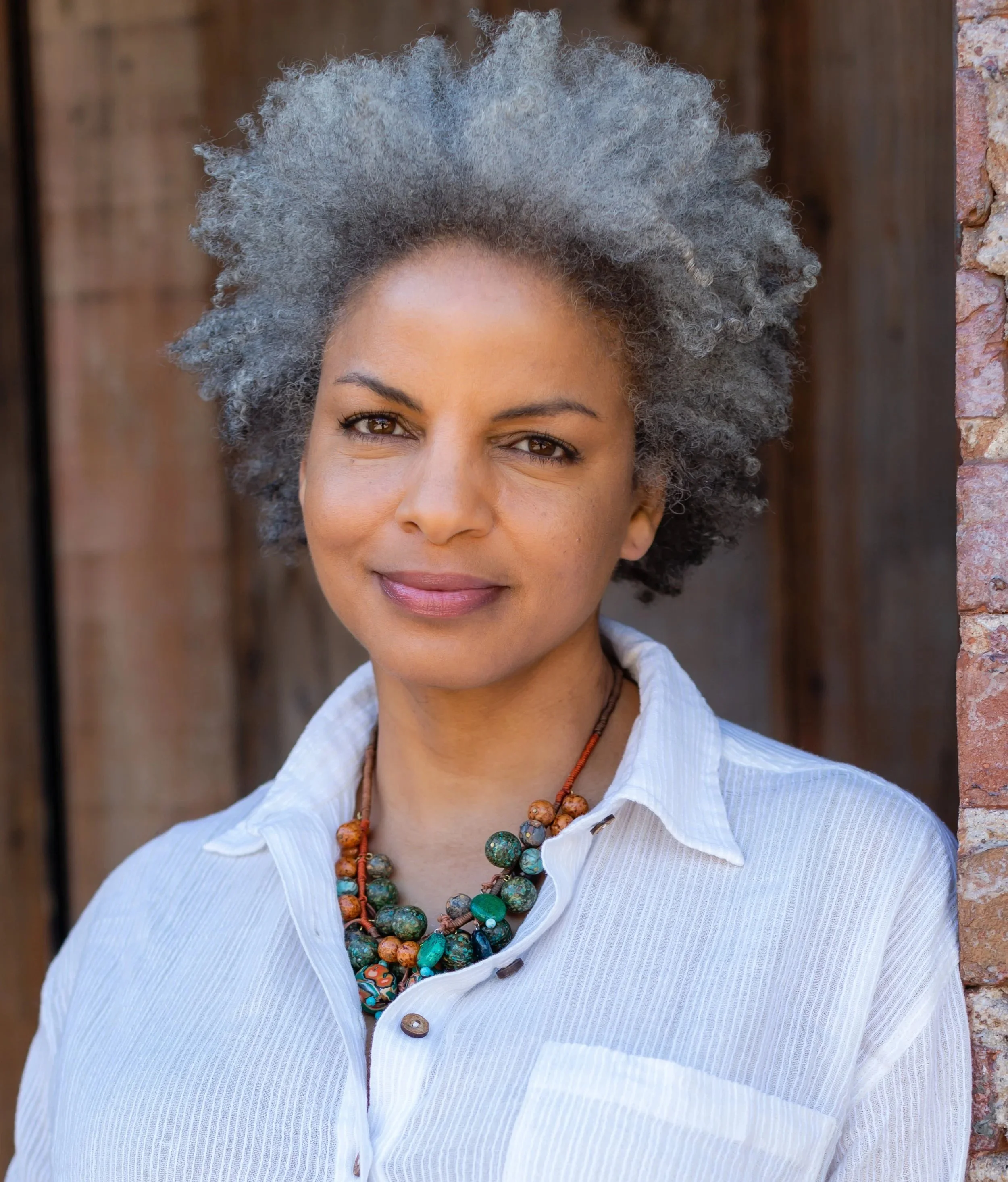 Close-up of a woman with gray, curly hair wearing a white shirt and colorful beaded necklace, standing outdoors near a brick wall and wooden background.